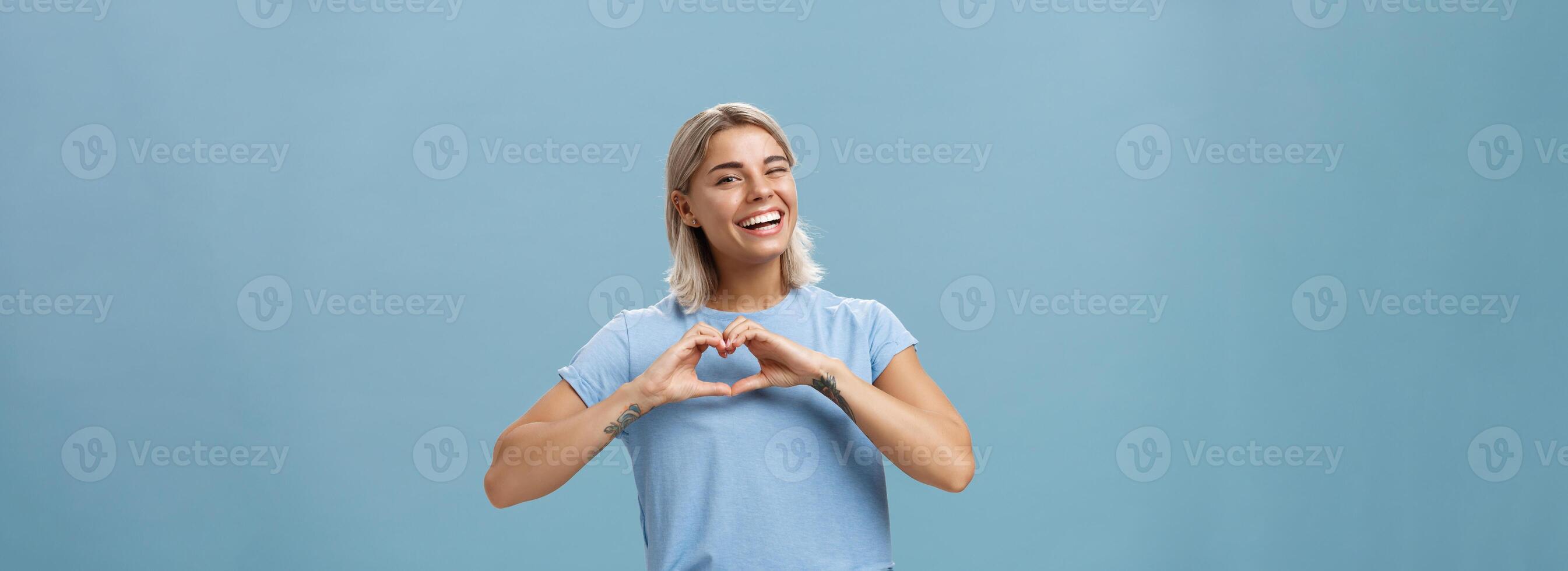 Love relationship and emotions concept. Joyful charming feminine girl with fair hair in trendy t-shirt winking happily smiling broadly and making heart gesture over breast expressing affection photo