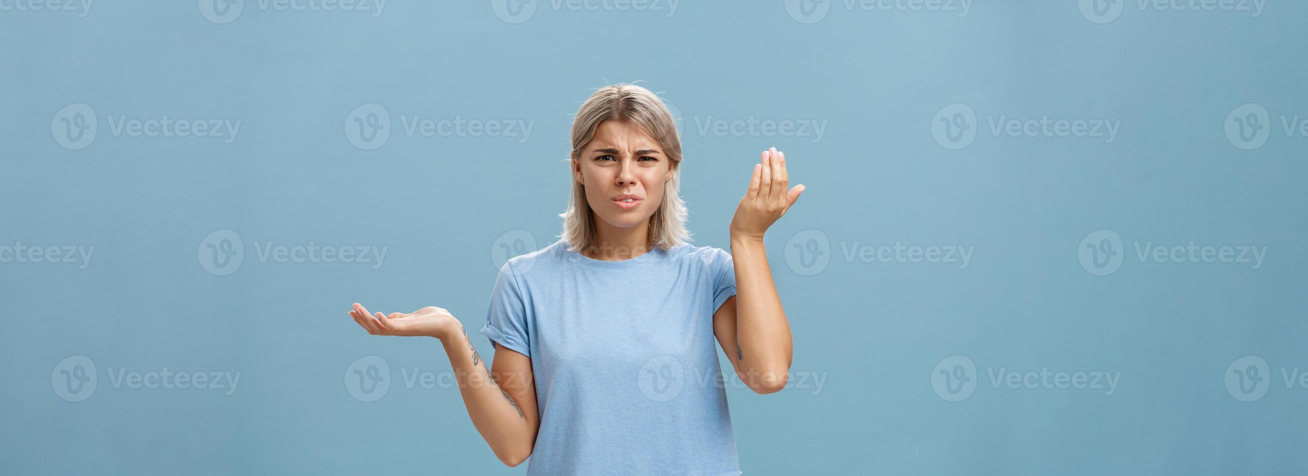 Wtf you saying. Portrait of pissed and confused stylish girlfriend with blond hair raising palm high and aside with perplexed expression being questioned hearing stupid nonsense over blue wall photo