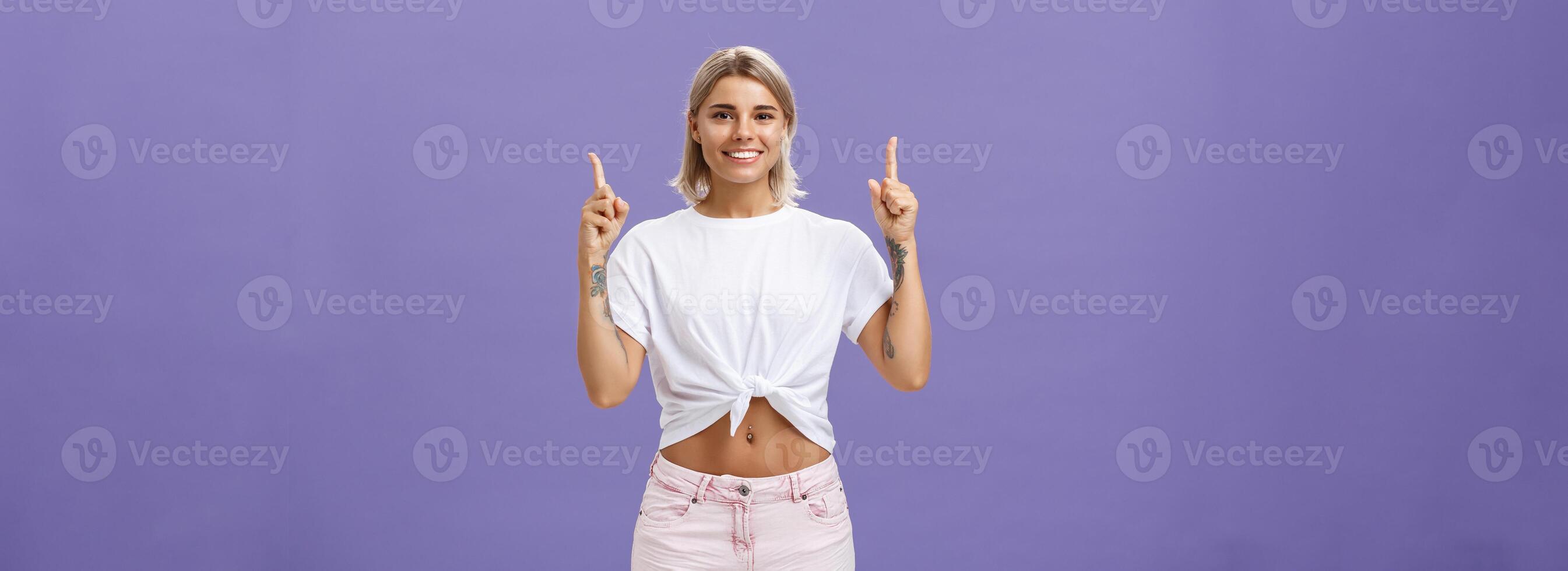 Time look upwards and move forward corporate ladder. Portrait of attractive ambitious and stylish young blonde woman with tattoos and pierced belly pointing up and smiling broadly over purple wall photo