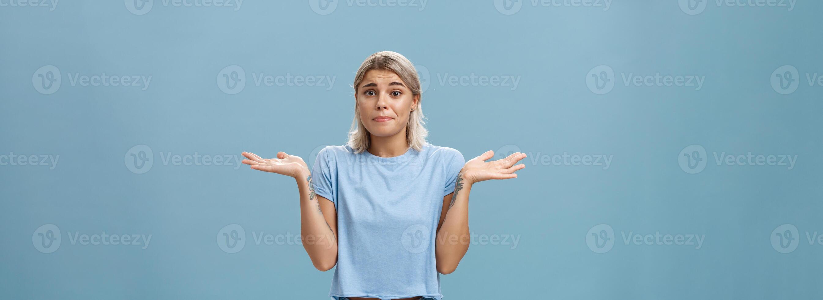 No idea how help sorry. Portrait of unsure gloomy girlfriend feeling regret shrugging with hands near shoulders frowning and pursing lips being uncertain and unaware over blue background photo