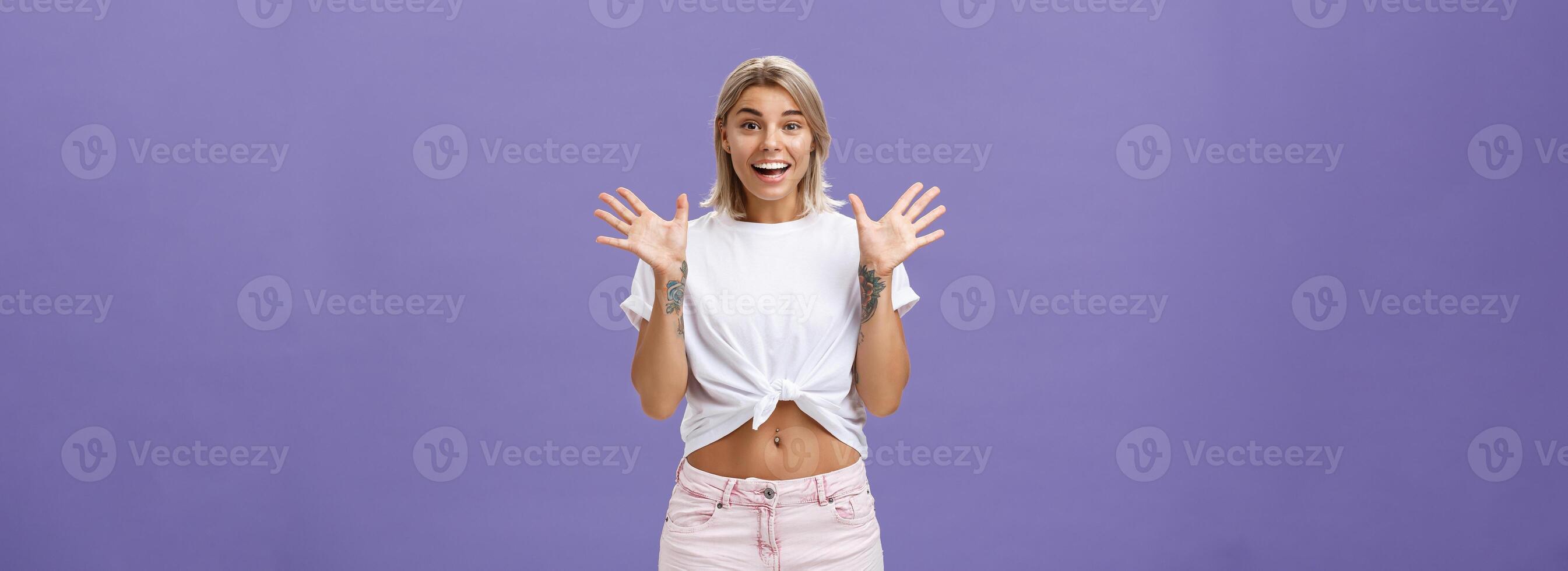 Indoor shot of delighted and amused attractive stylish urban female with blond hair in stylish white t-shirt and summer shorts raising palms from surprise and amazement standing over purple background photo