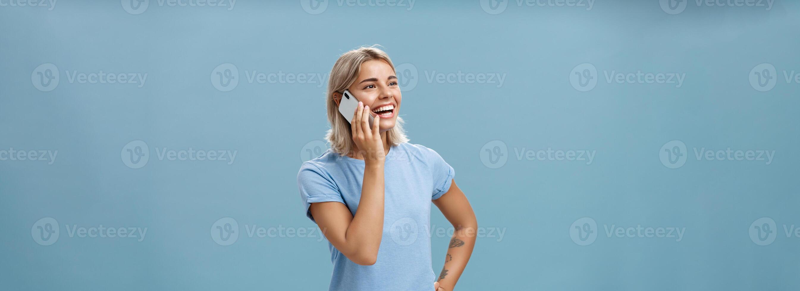 Waist-up shot of sociable amused and happy attractive caucasian fair-haired woman in casual t-shirt standing half-turned gazing left with hand on hip while talking on smartphone over blue background photo