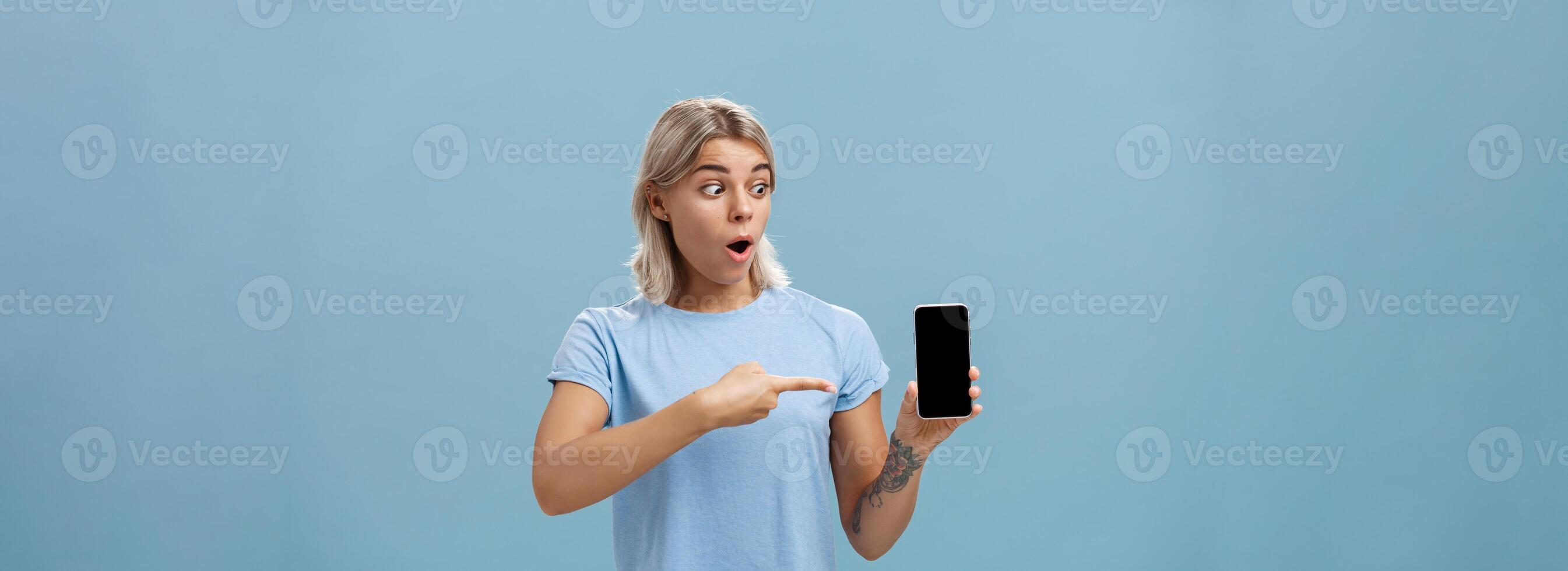 Girl being amazed with cool new smartphone received on birthday. Surprised speechless happy woman with fair hair and tattoo dropping jaw holding smartphone pointing and staring at gadget screen photo