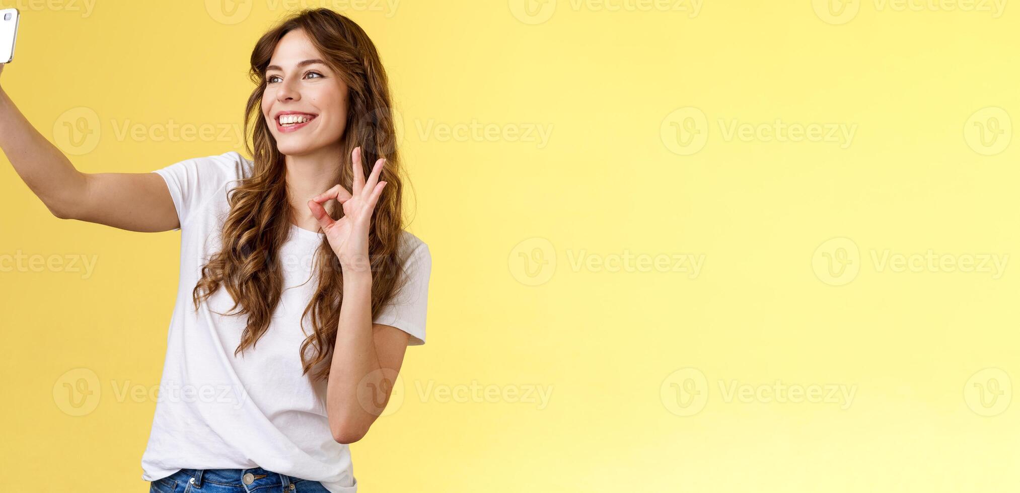 Stylish carefree urban confident curly-haired girl recording video blog followers extend arm hold smartphone taking selfie front camera show okay ok approval gesture yellow background photo