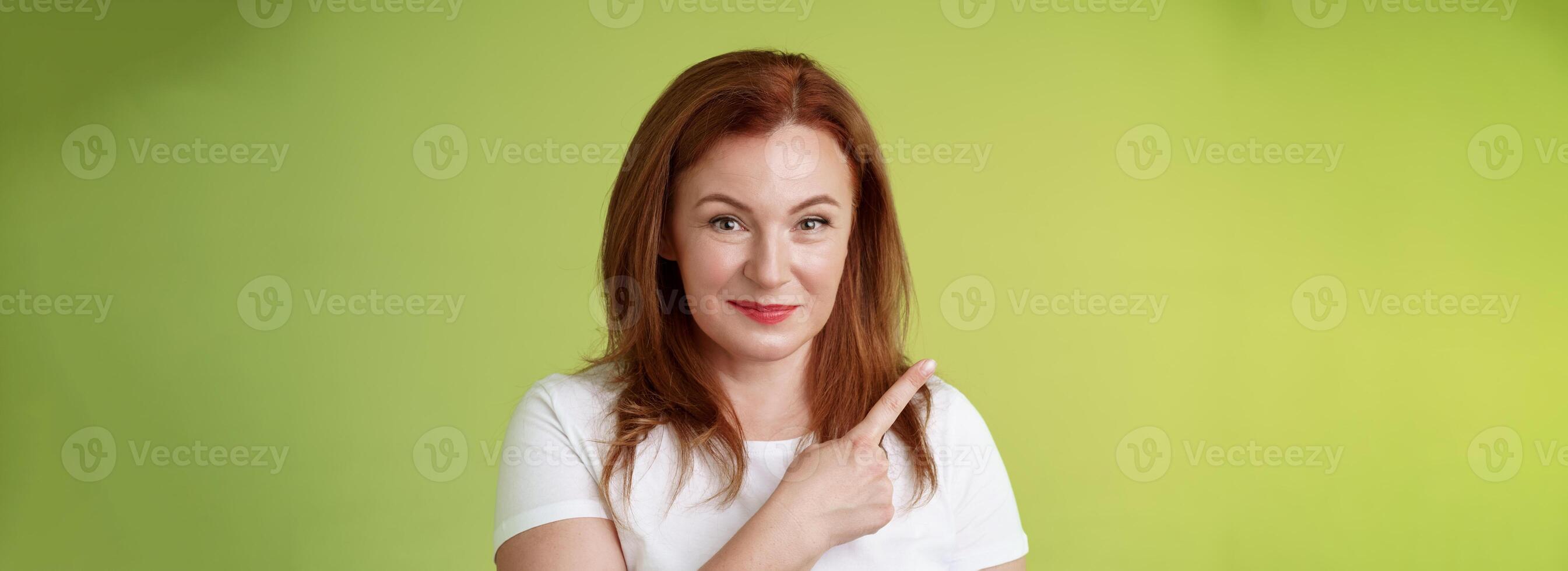 Kind cheerful good-looking middle-aged woman 50s redhead white t-shirt smiling modest assured give advice pointing upper left corner indicating great promotion advertising product green background photo