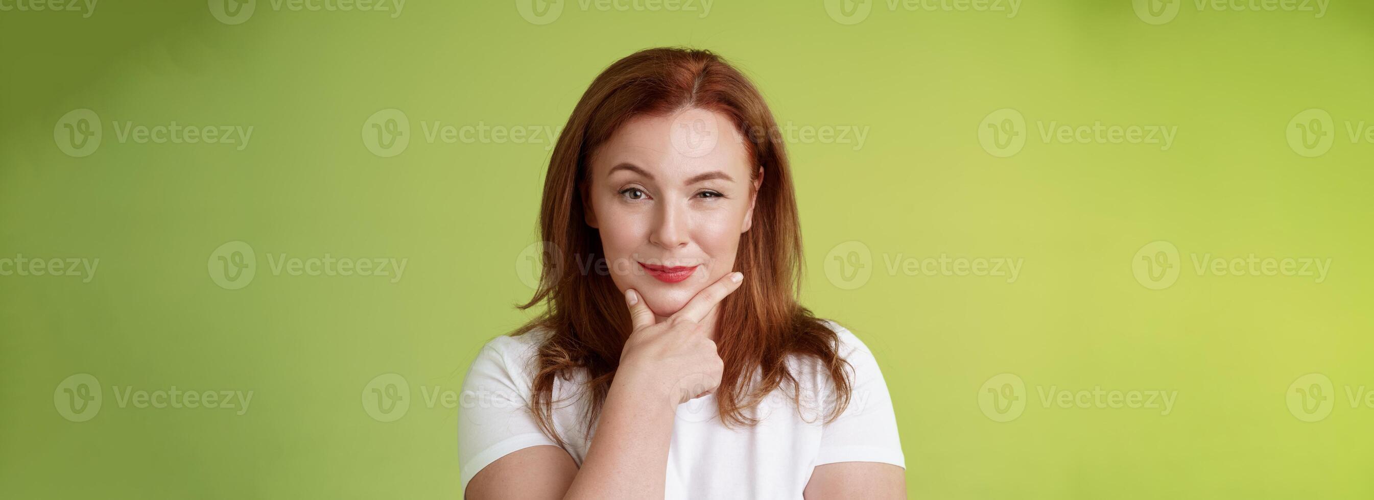 Hmm interesting choice. Intrigued cunning redhead middle-aged female hold hand face-line rub chin thoughtful smirking pleased raise eyebrow curious pondering alluring decision green background photo