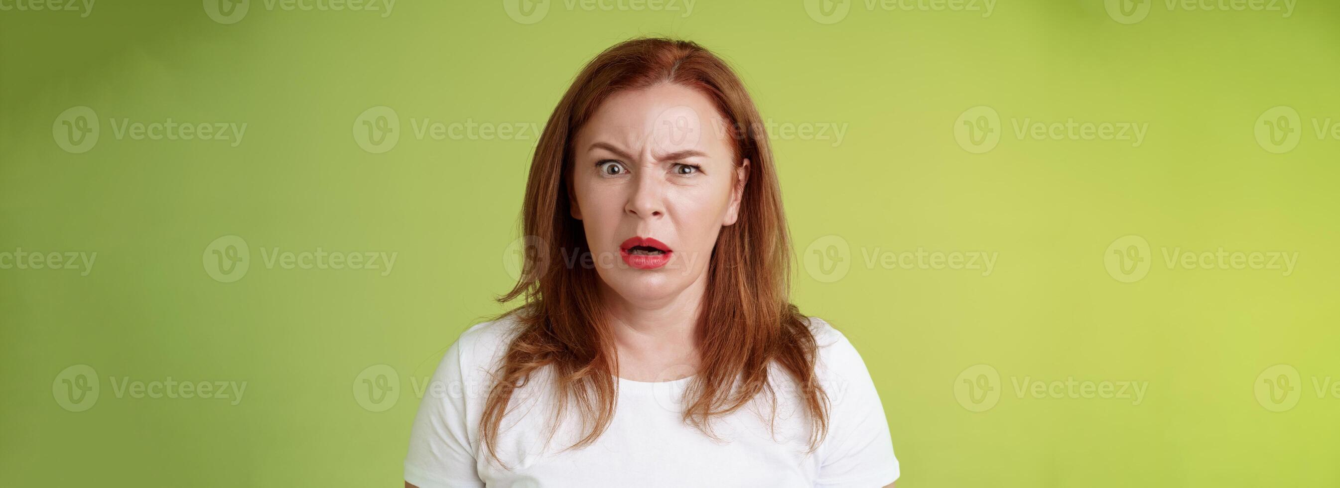 Confused shocked gasping middle-aged redhead woman cringe frustrated puzzled open mouth speechless freak out strange shocking scene stand green background perplexed disappointed photo