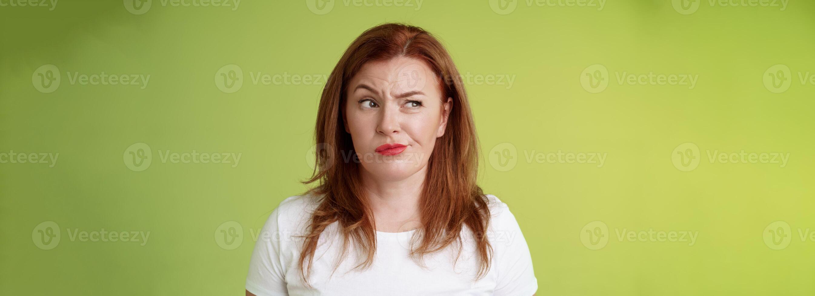 Perplexed doubtful unsure redhead wife smirking unsatisfied frowning look aside displeased disappointed hesitating pondering strange advice thoughtfully stare away stand green background photo