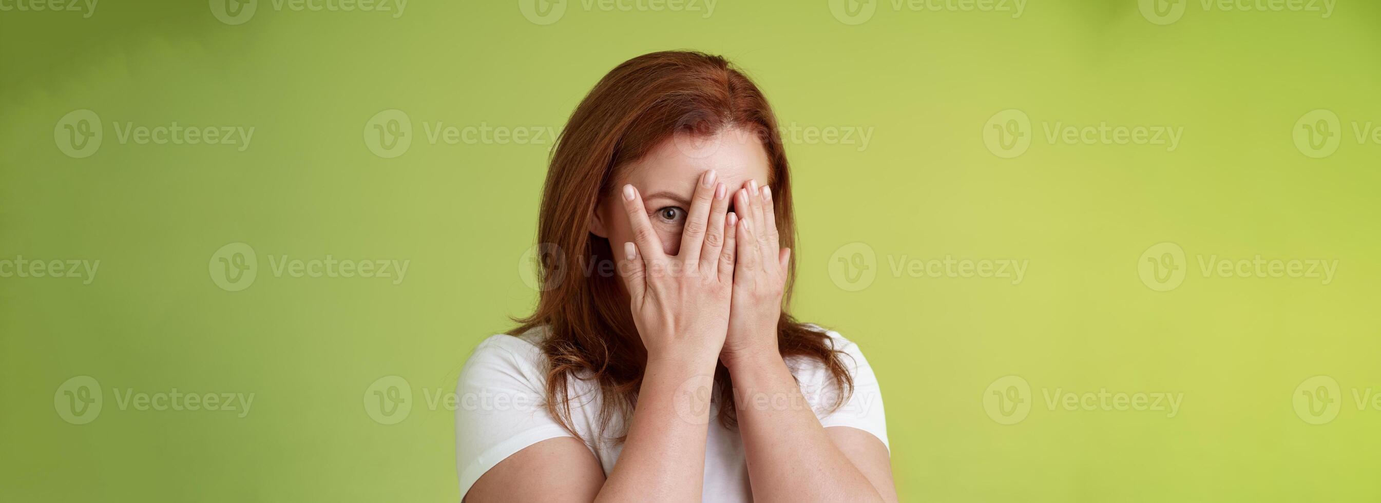 Not peeking. Playful charismatic middle-aged ginger redhead woman close face palms look through fingers one eye stare camera intrigued waiting surprise gift stand green background photo