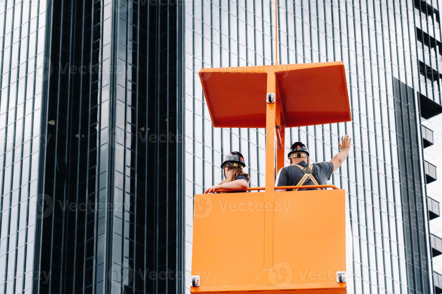 Workers in a construction cradle climb on a crane to a large glass building.The crane lifts the workers in the car seat.Construction photo