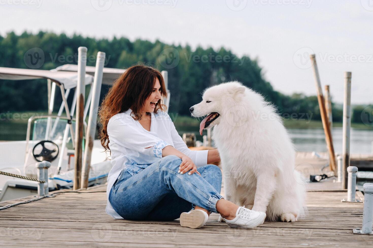 a happy woman with a big white dog sits on a pier by the sea at sunset photo