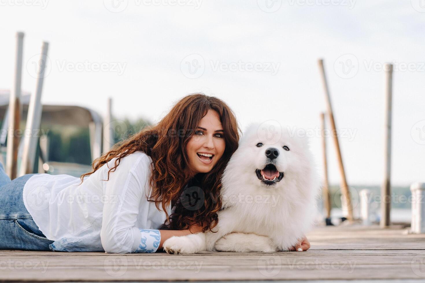 un contento mujer con un grande blanco perro mentiras en un muelle cerca el mar a puesta de sol foto