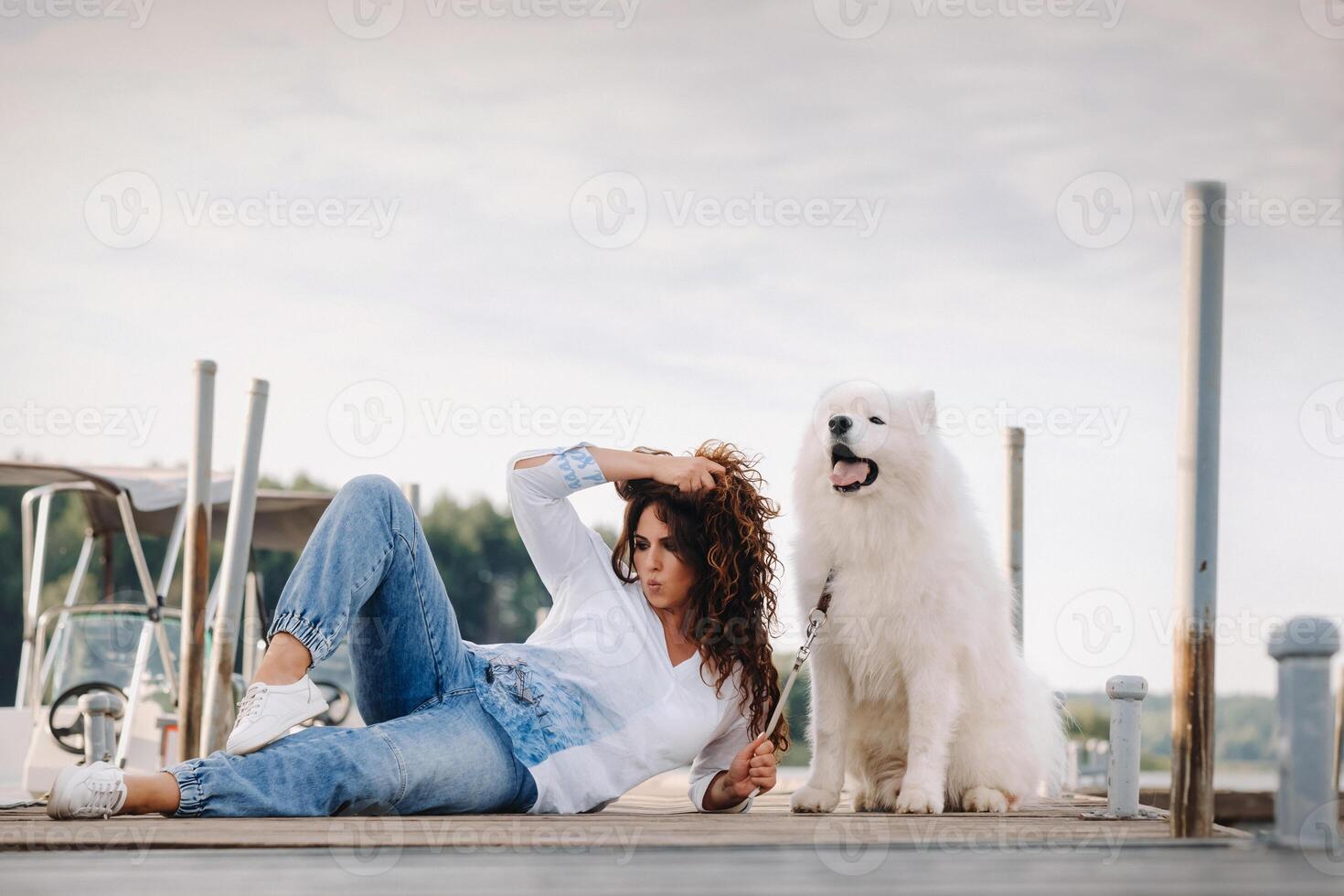 un contento mujer con un grande blanco perro mentiras en un muelle cerca el mar a puesta de sol foto