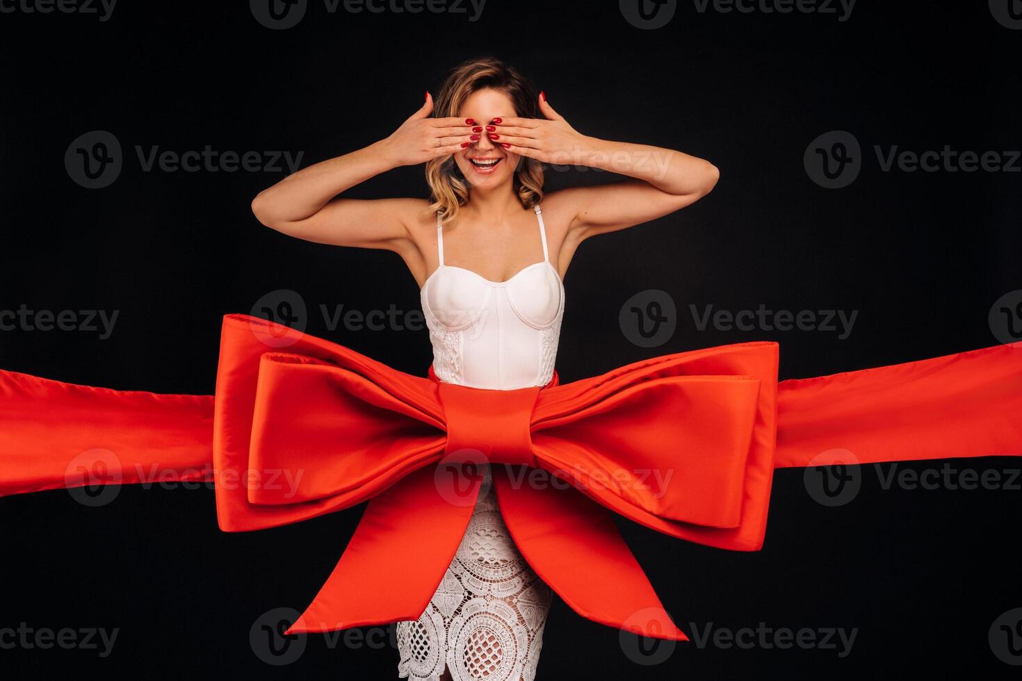 a woman in a white dress as a gift covers her eyes with her hands on a black background wrapped in a festive ribbon photo