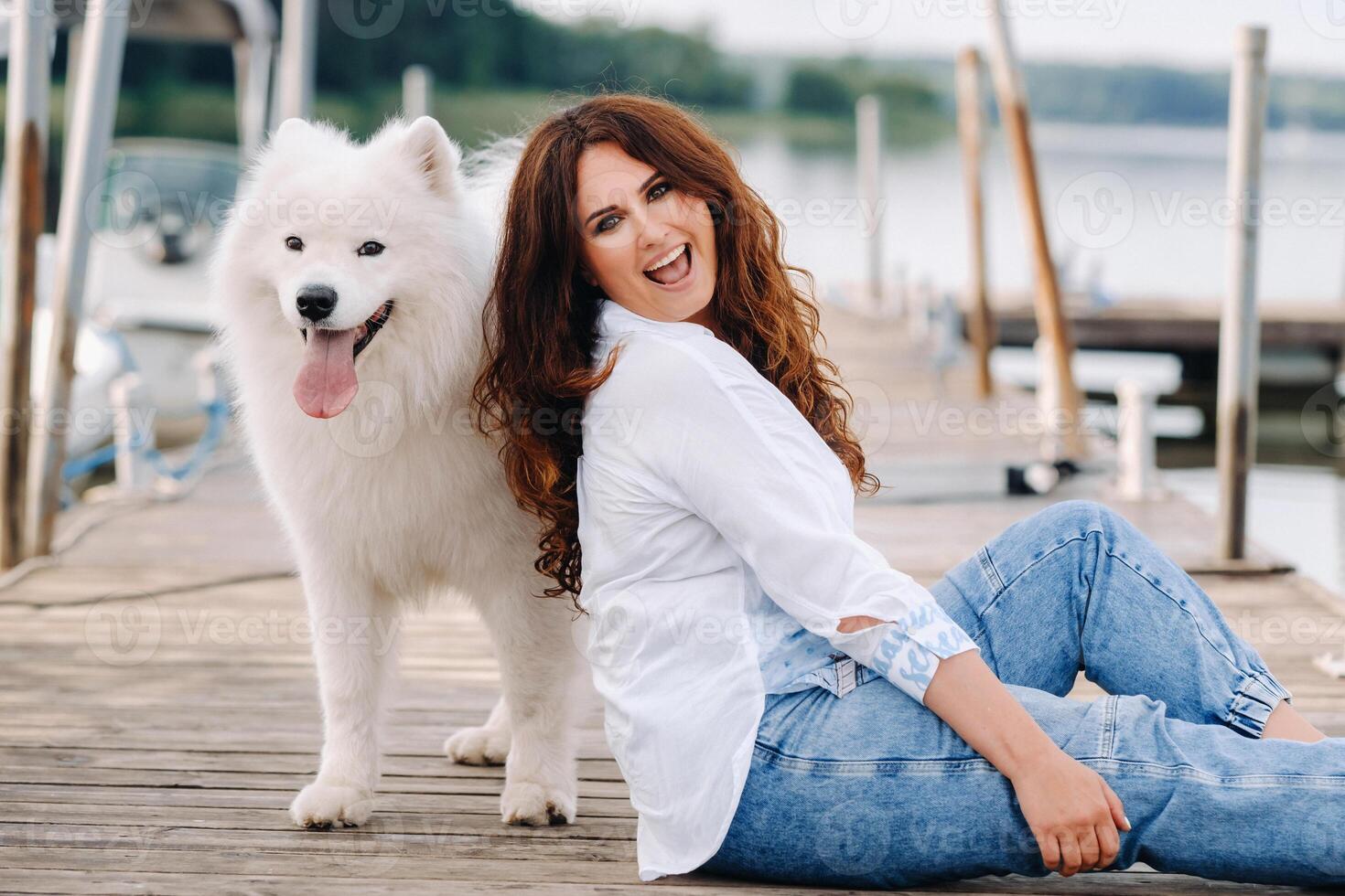 un contento mujer con un grande blanco perro se sienta en un muelle por el mar a puesta de sol foto