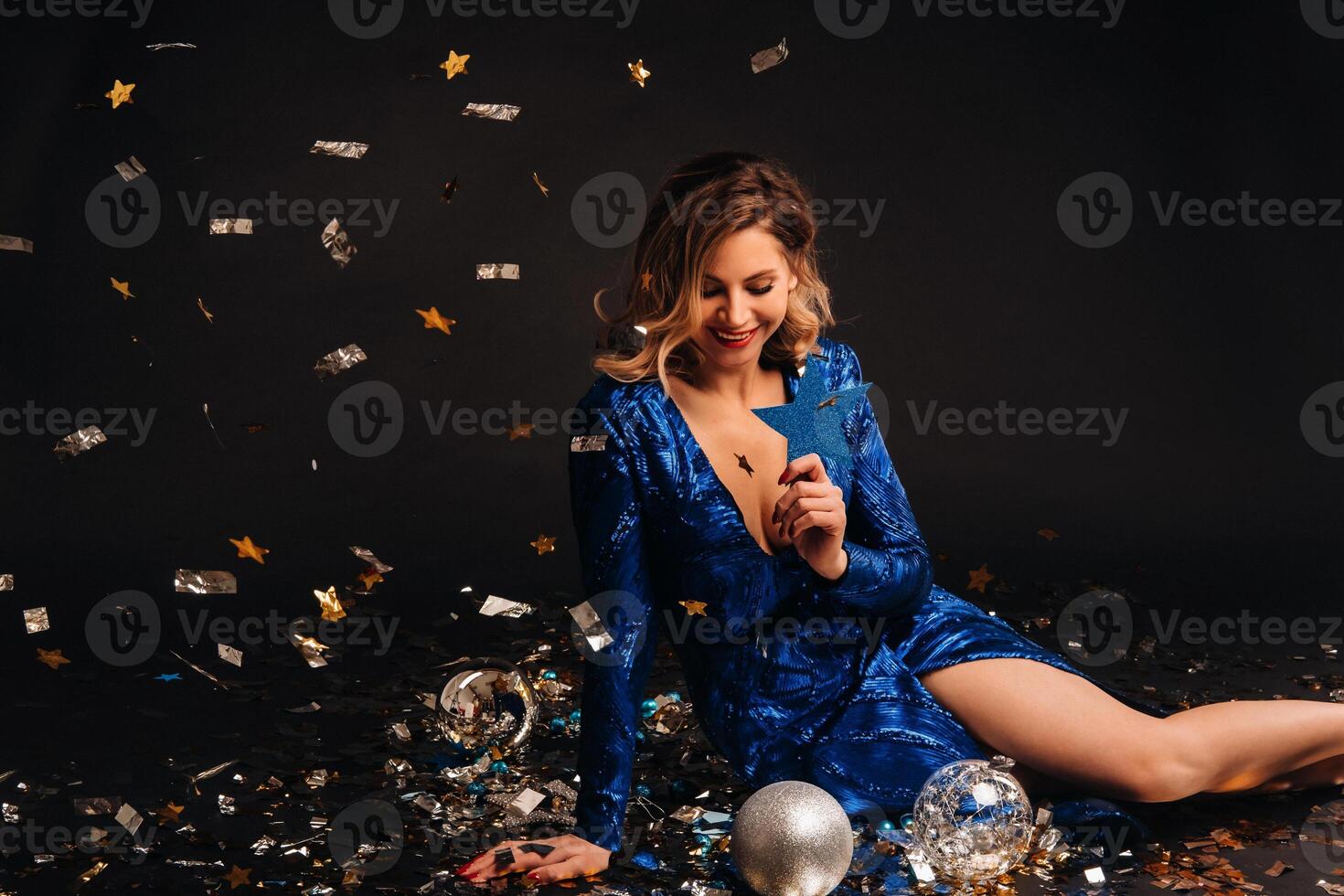 a woman in a blue sequined dress is smiling sitting on the floor with confetti on a black background photo