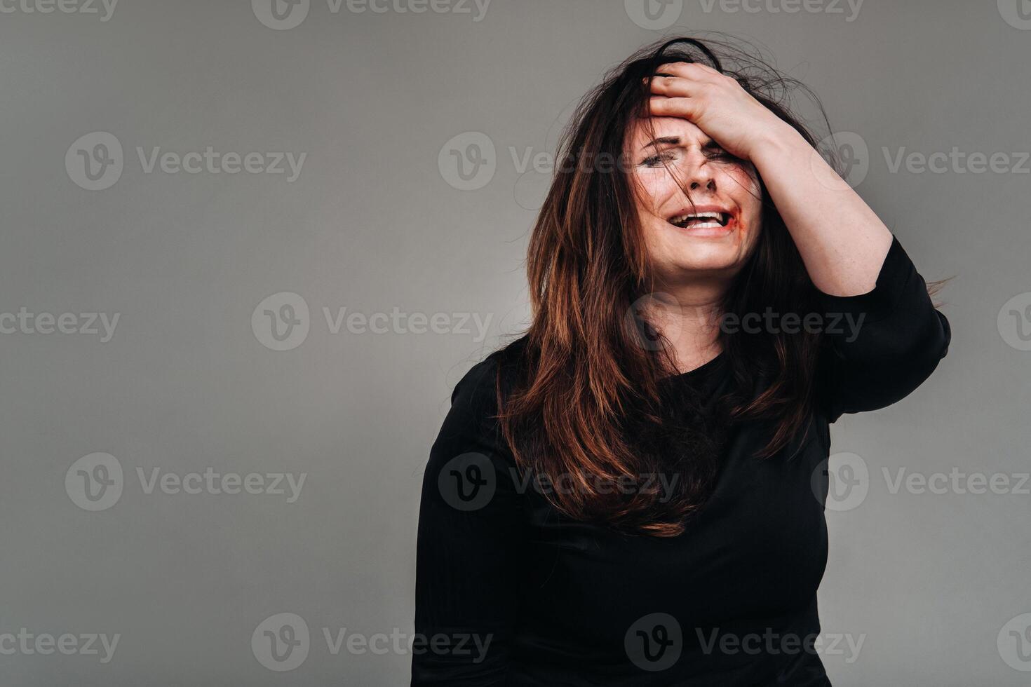 a battered woman in black clothes holds her head against an isolated gray background. Violence against women photo