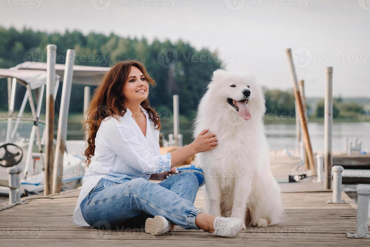 un contento mujer con un grande blanco perro mentiras en un muelle cerca el mar a puesta de sol foto