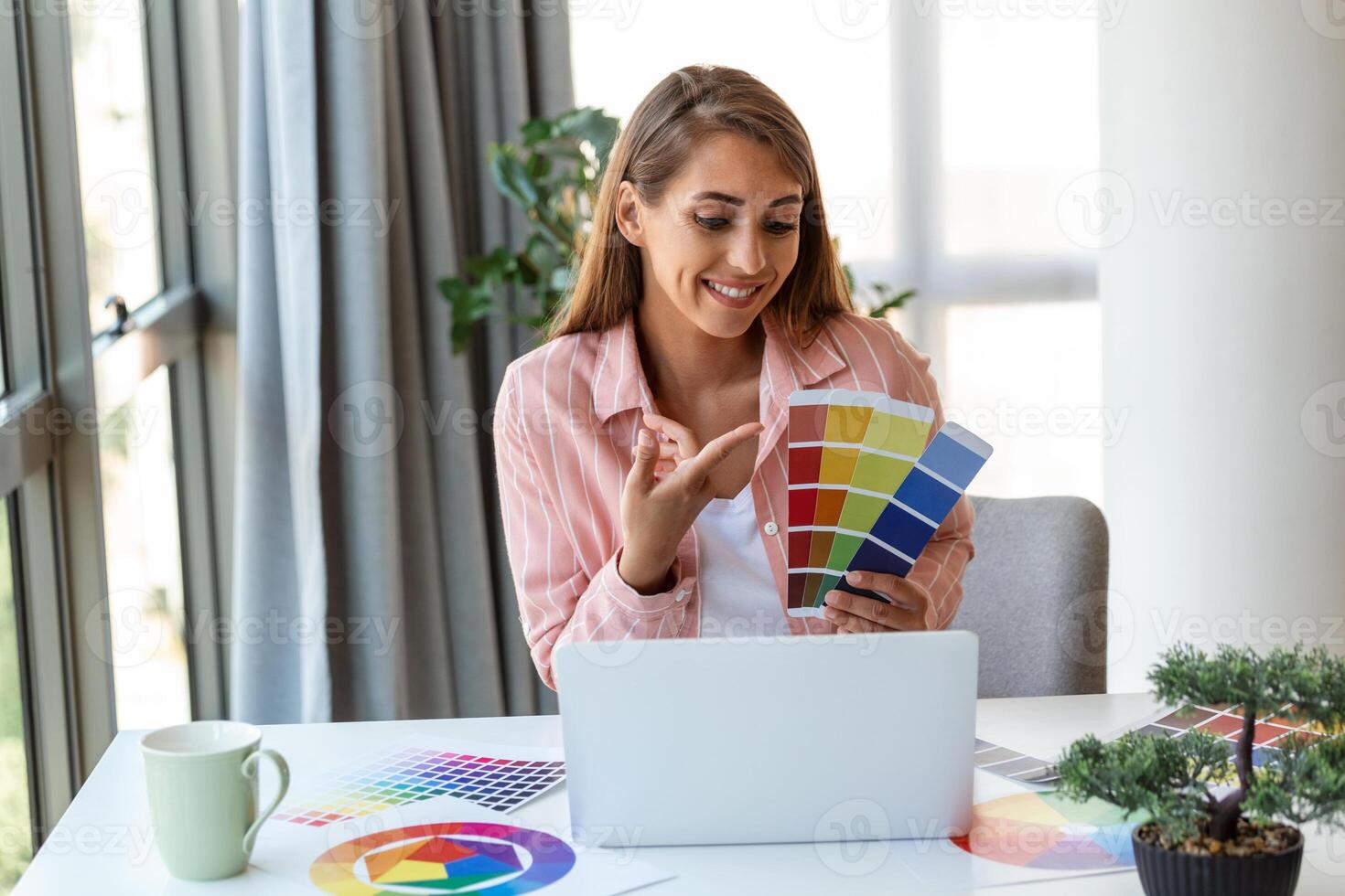 Cheerful young woman designer having video conference with clients, sitting at desk in front of computer, holding color palettes, gesturing and smiling, copy space photo