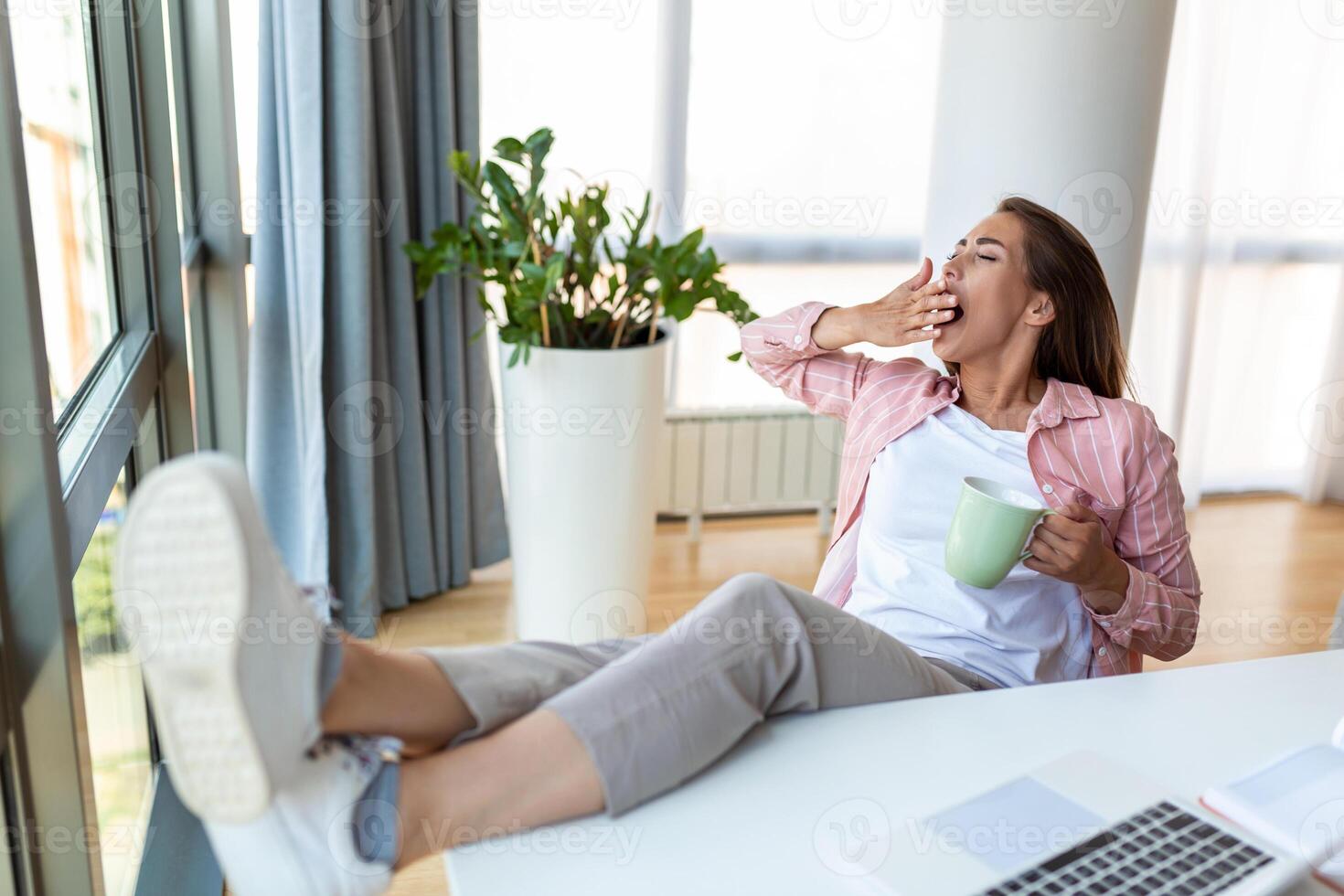 Young pretty businesswoman in the office yawning sitting in the chair, hands covering her mouth with her feet up on a desk drinking coffee sleepy photo