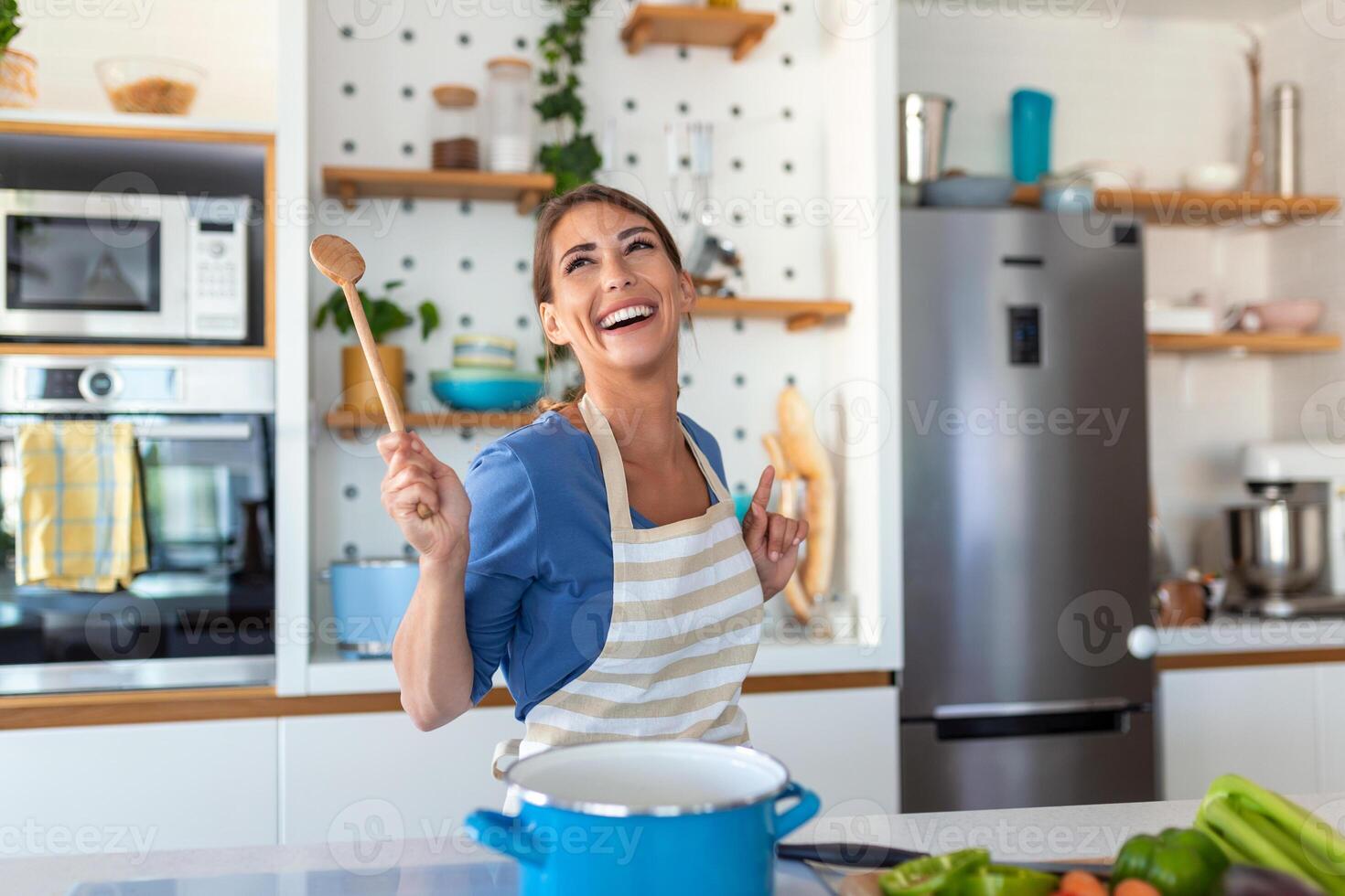 emocionado mujer canto y bailando en moderno cocina a hogar, contento mujer participación espátula como micrófono, baile, escuchando a música, teniendo divertido con batería de cocina, preparando desayuno foto