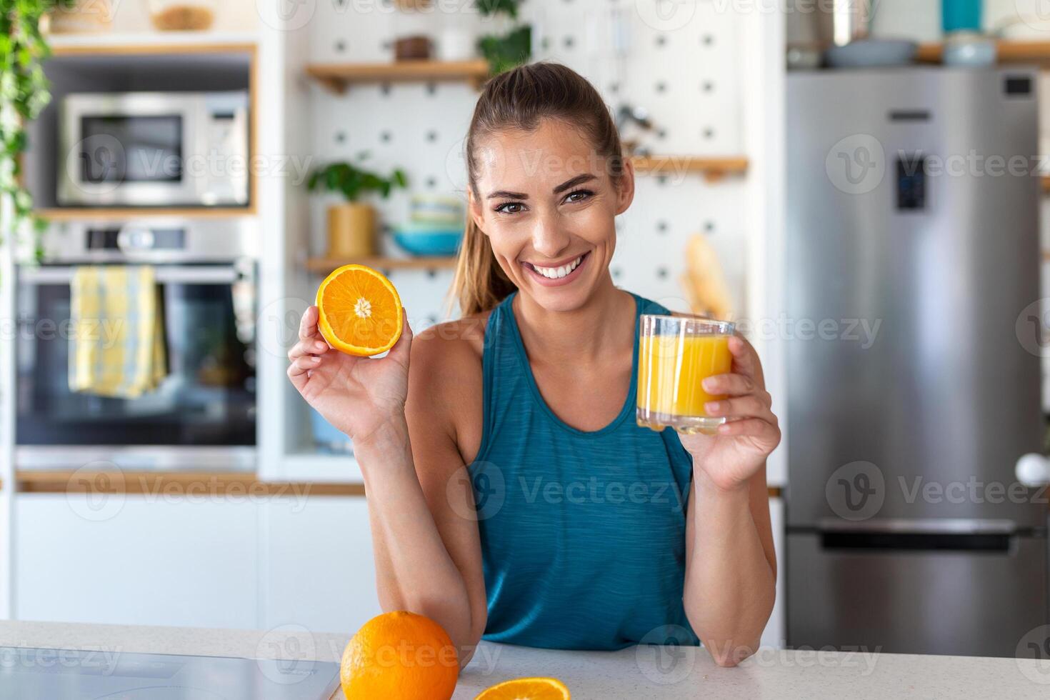 Beautiful young woman drinking fresh orange juice in kitchen. Healthy diet. Happy young woman with glass of juice and orange at table in kitchen. photo