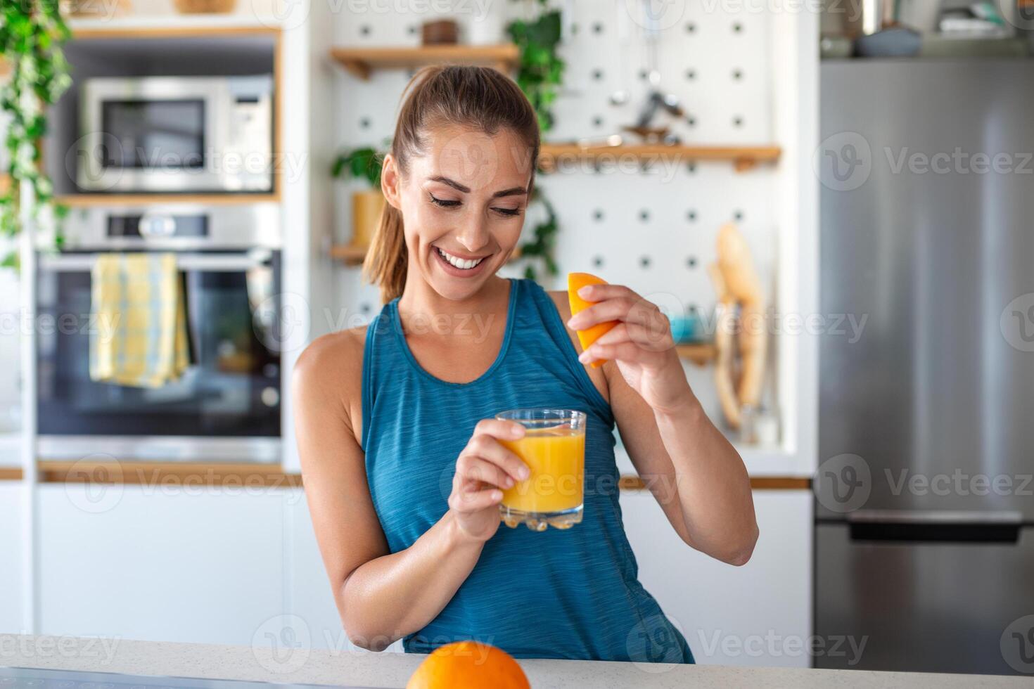 Beautiful young woman drinking fresh orange juice in kitchen. Healthy diet. Happy young woman with glass of juice and orange at table in kitchen. photo