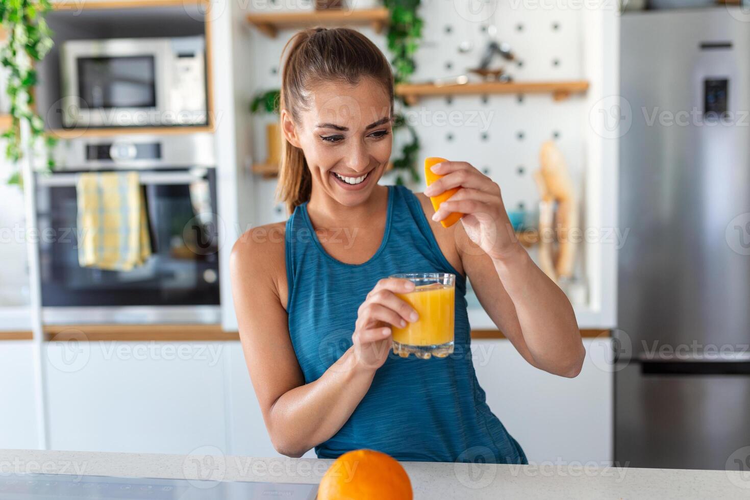 Beautiful young woman drinking fresh orange juice in kitchen. Healthy diet. Happy young woman with glass of juice and orange at table in kitchen. photo