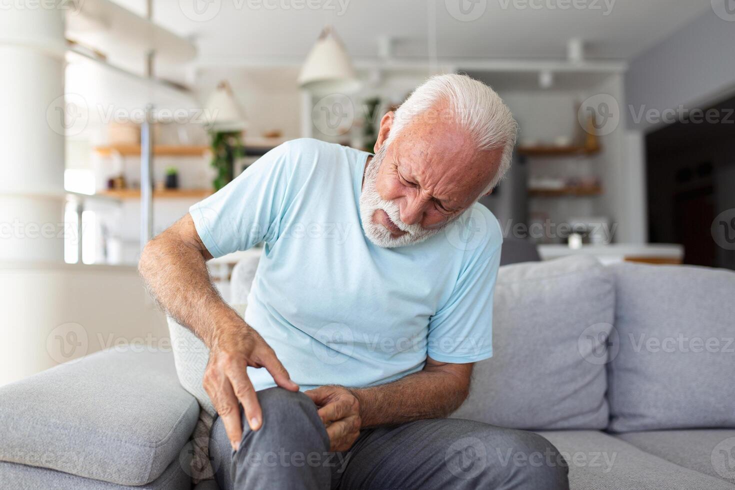 The older man is sitting on the couch at home, has pain in the knee joint, holding his leg, osteoarthritis concept. photo