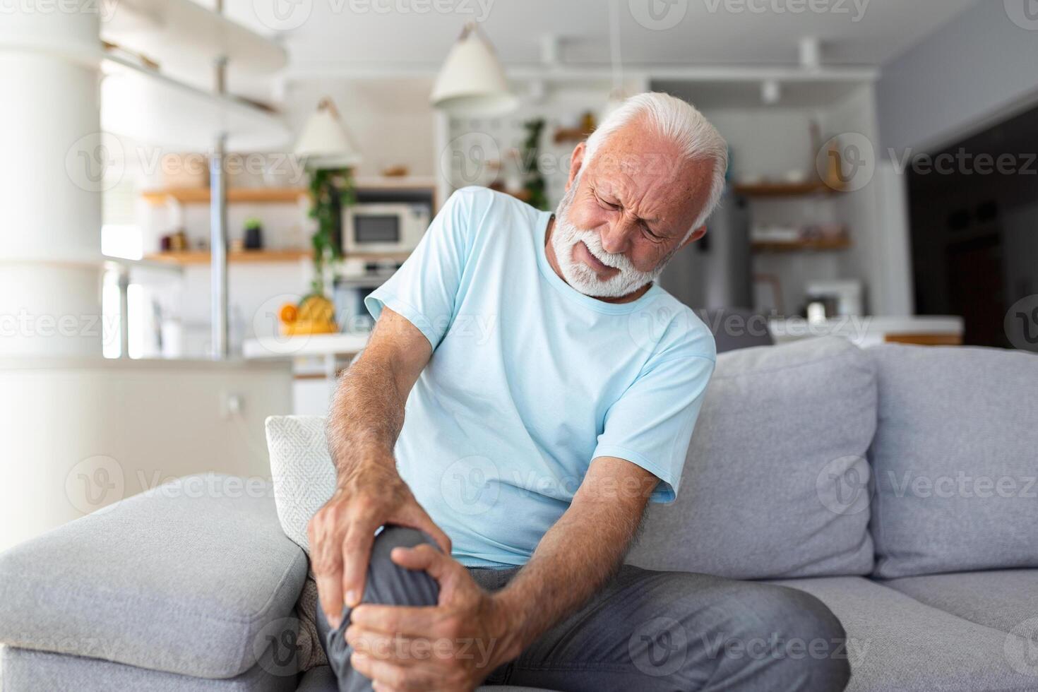 The older man is sitting on the couch at home, has pain in the knee joint, holding his leg, osteoarthritis concept. photo