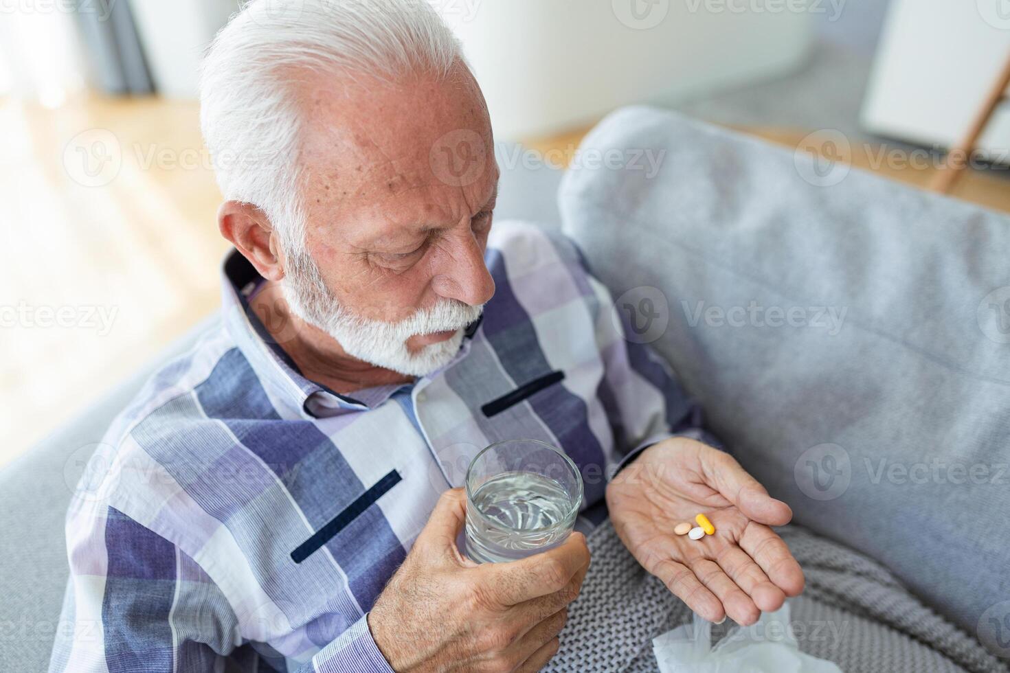 Senior man taking pills while sitting on couch at home, holding white jar with treatment, copy space. Grey-haired elderly man using supplements or vitamins. Healthy lifestyle in senior age concept photo