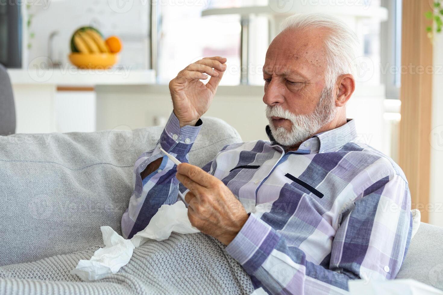 Sick elderly man checking his temperature suffering from seasonal flu or cold lying on sofa caughing suffering from seasonal flu or cold. . Ill senior feel unhealthy with influenza at home photo