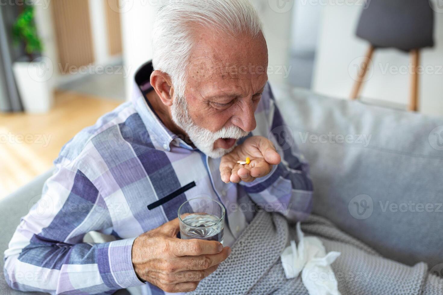 Senior man taking pills while sitting on couch at home, holding white jar with treatment, copy space. Grey-haired elderly man using supplements or vitamins. Healthy lifestyle in senior age concept photo