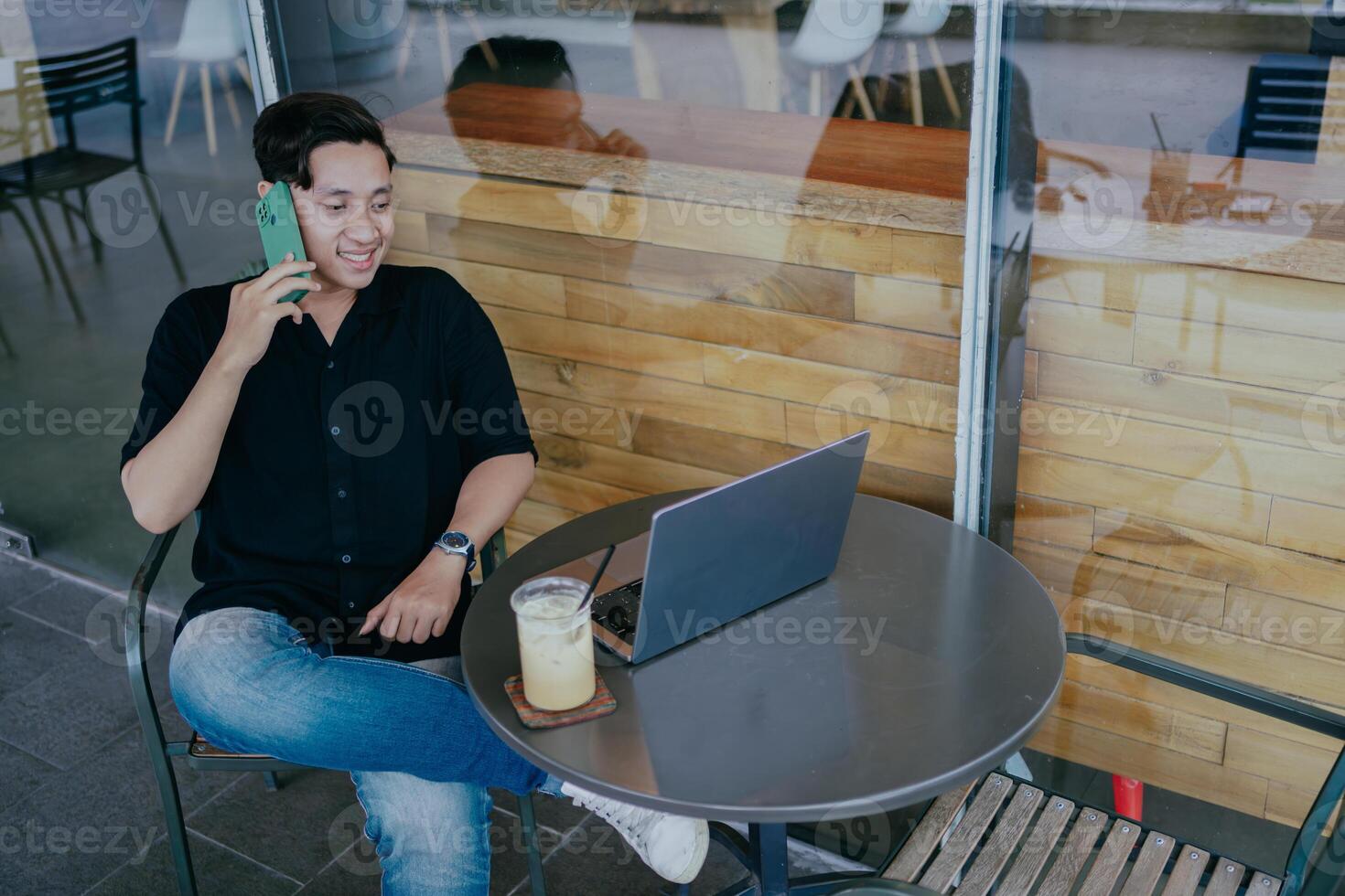 sonriente joven masculino empresario hablando en el teléfono, sentado relajado a café mesa con ordenador portátil y vaso de con hielo Leche café. contento hombre chateando con amigos distraído desde trabajar. foto