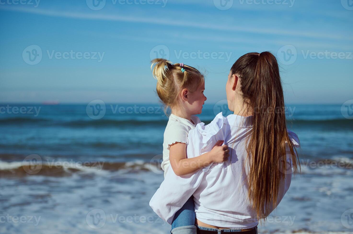 Rear view of mother holding her daughter and admiring waves splashing while pounding on the sandy shore. Family pastime photo