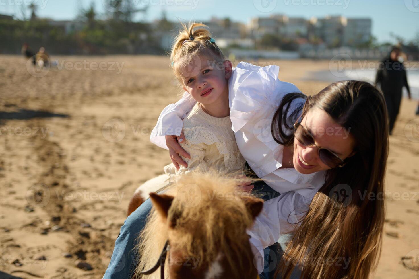 Smiling mother stroking a little pony while supports her daughter on horse riding. People. Active healthy lifestyle. photo