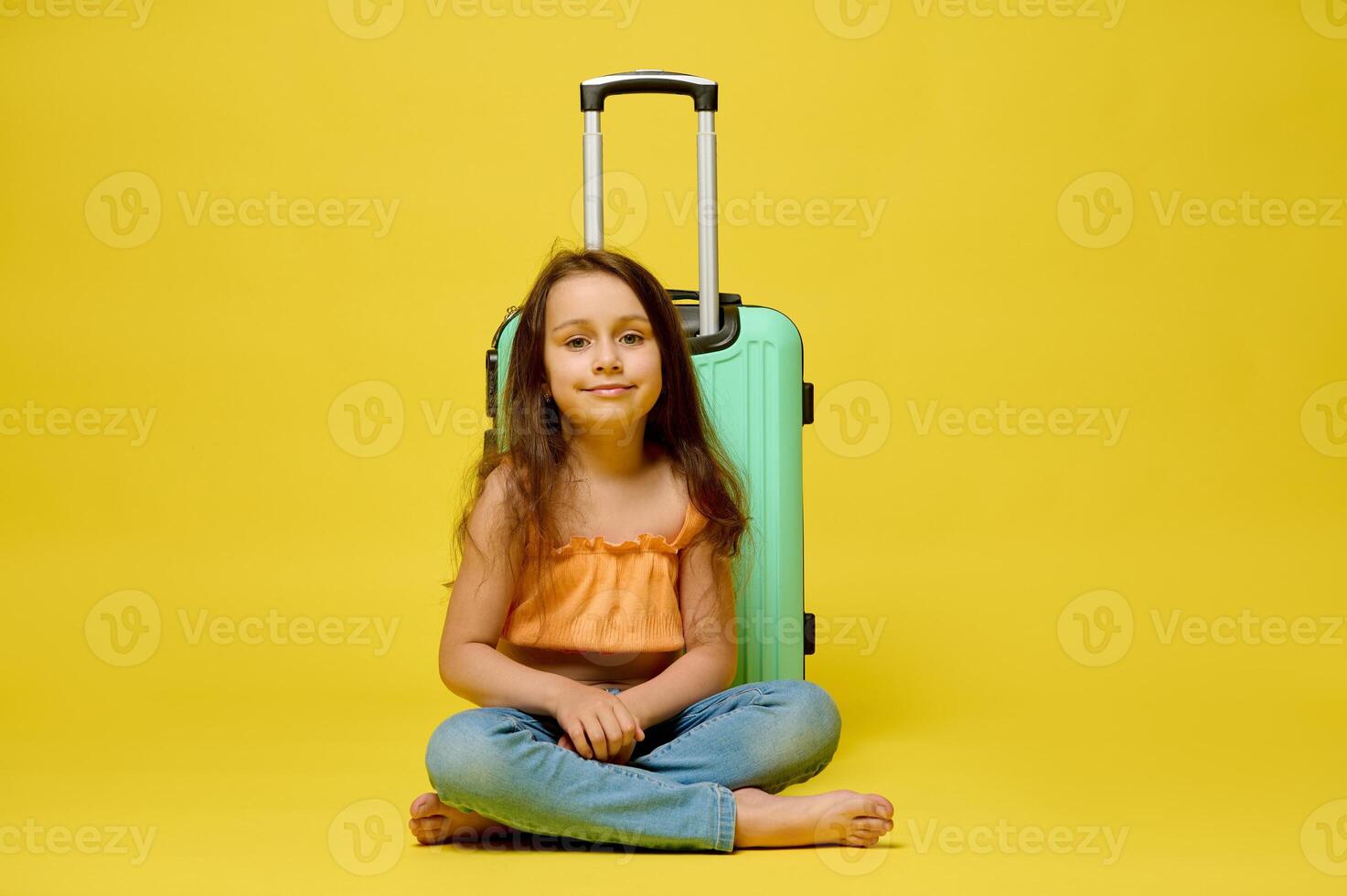 Caucasian cute traveler child girl with a suitcase, sitting over yellow studio background, smiling, looking at camera. photo