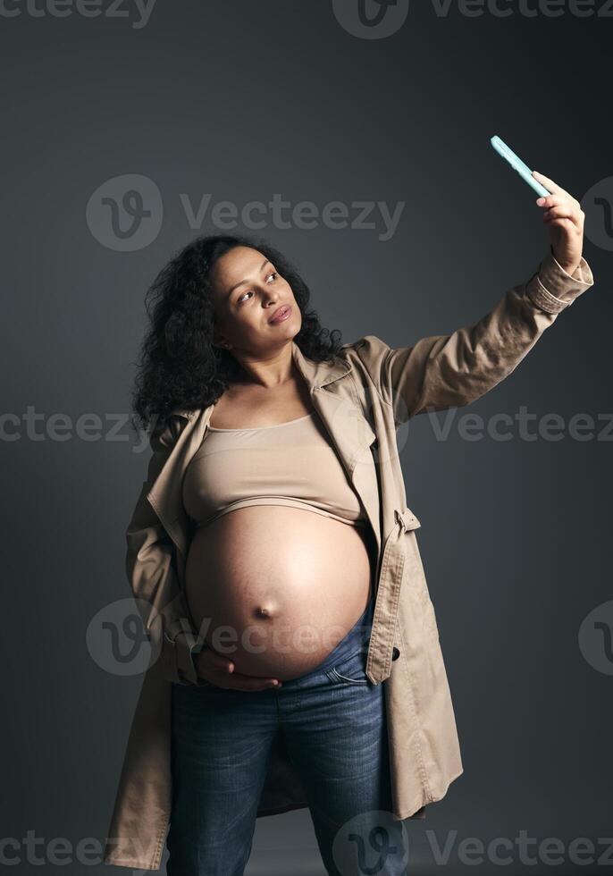 Happy pregnant woman takes self portrait on mobile phone while strokes her big belly, isolated fashion gray background photo
