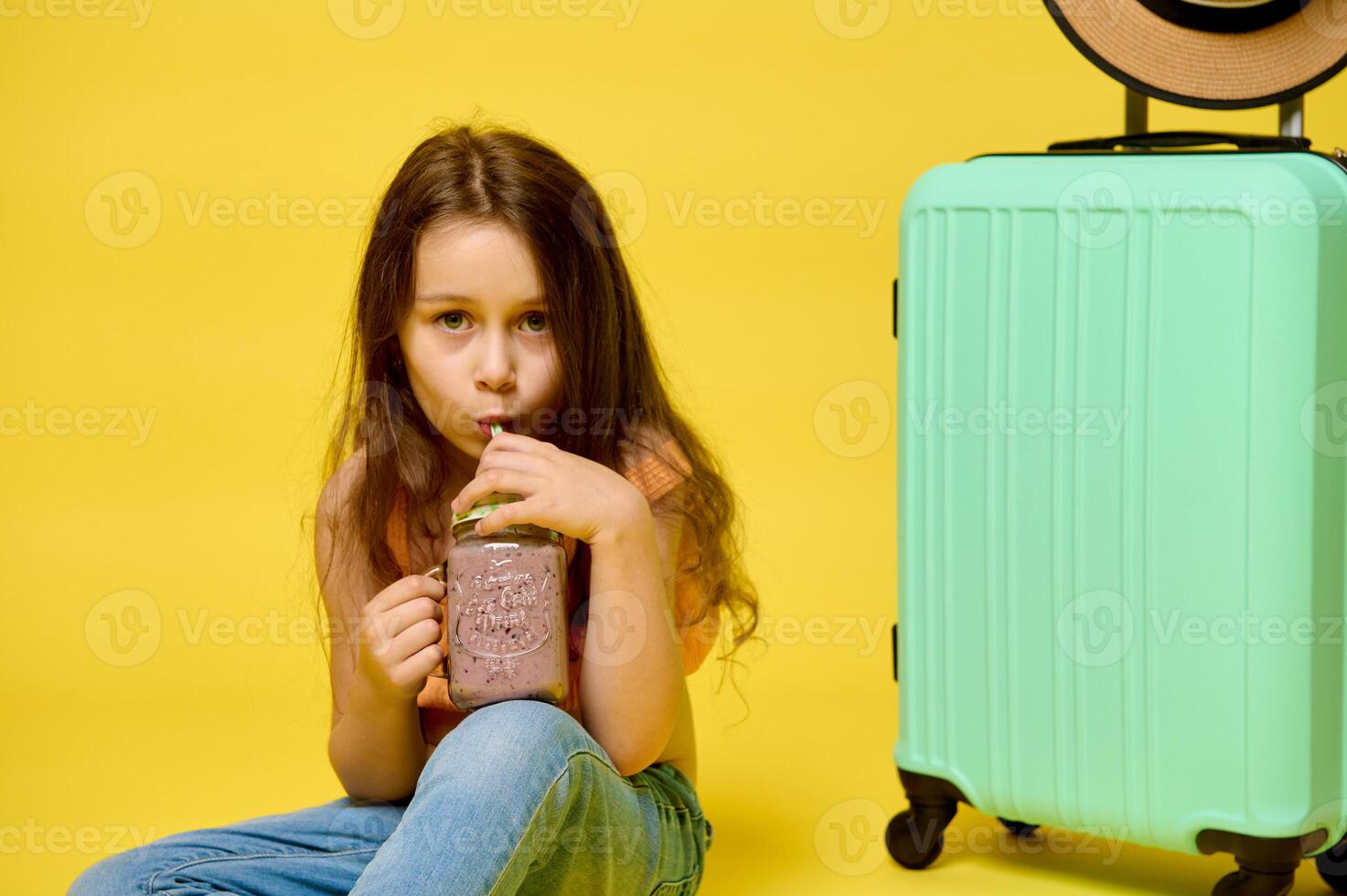Adorable child girl drinking a fresh cocktail from straw, sitting near suitcase, isolated over yellow studio background. photo