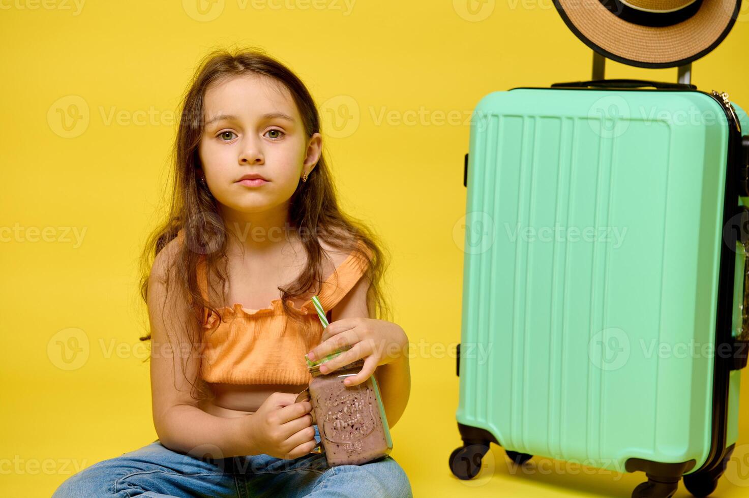 Lovely little kid girl drinking a cocktail, sitting near a trendy suitcase, looking at camera, isolated yellow backdrop photo