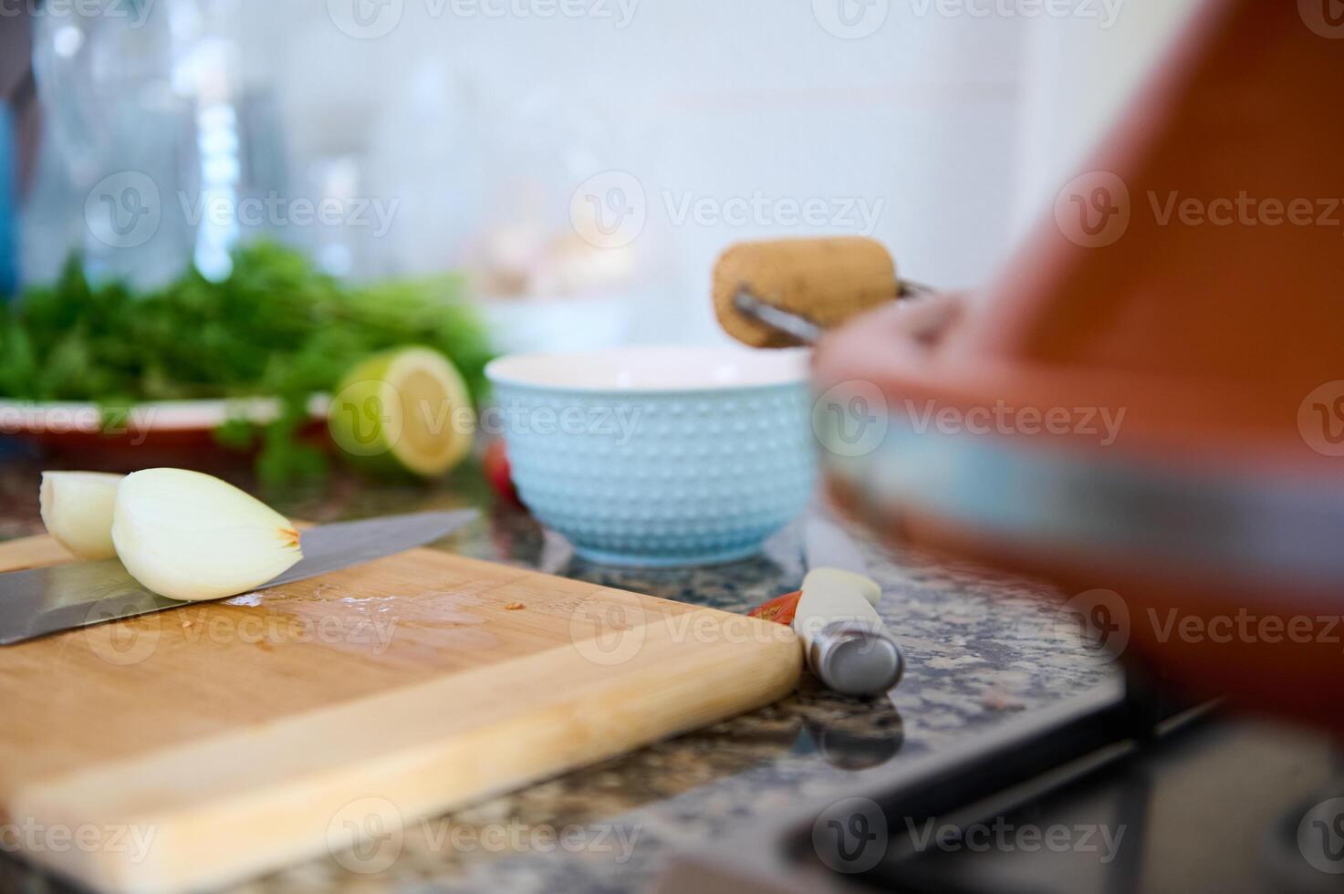 Cropped view of halves of an onion on a wooden board on the kitchen counter and a clay pot tagine steaming on the stove photo