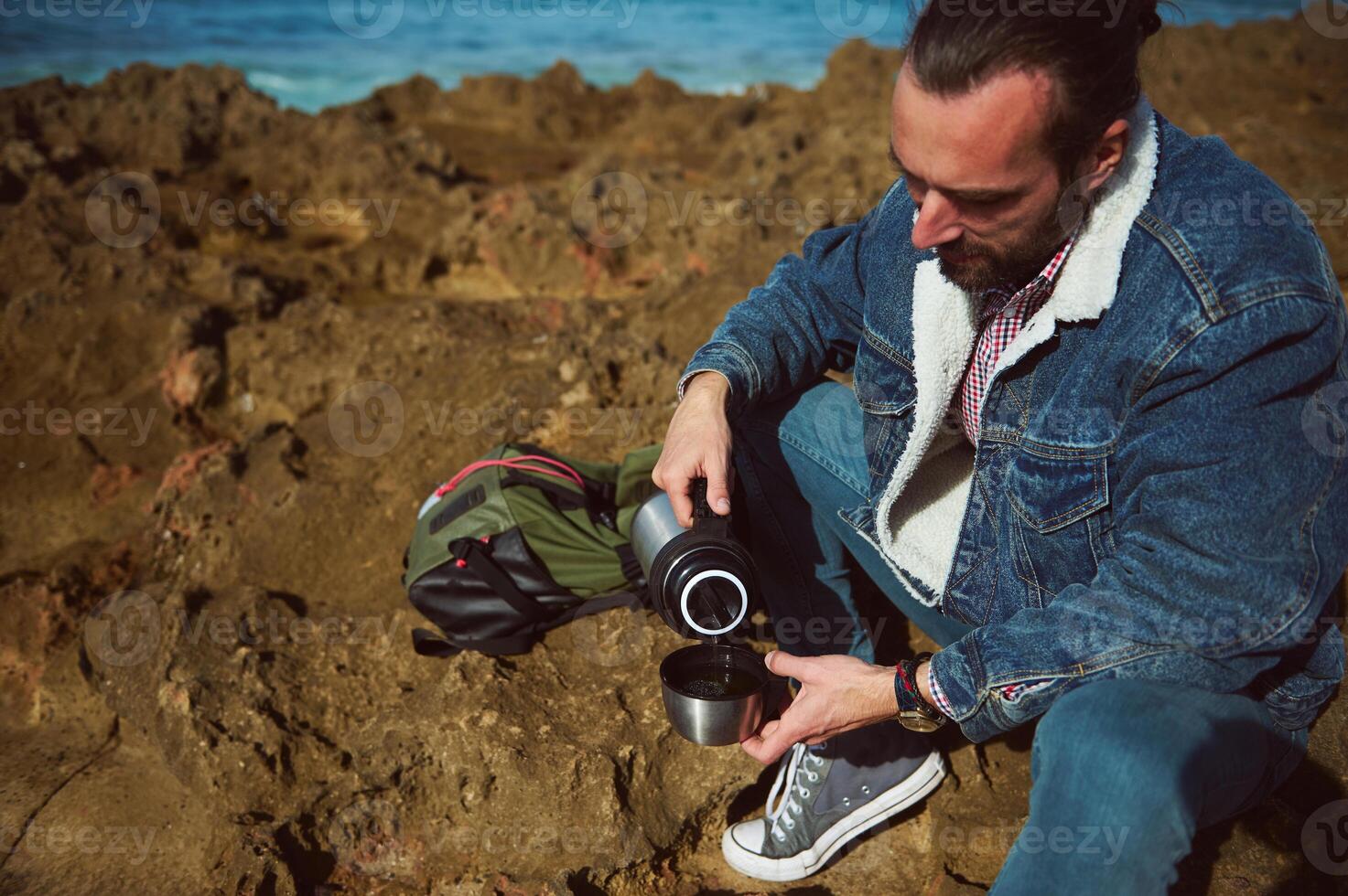 Relaxed young male tourist sitting on a rock by sea, holding a thermos bottle and pouring some coffee into a steel mug. photo
