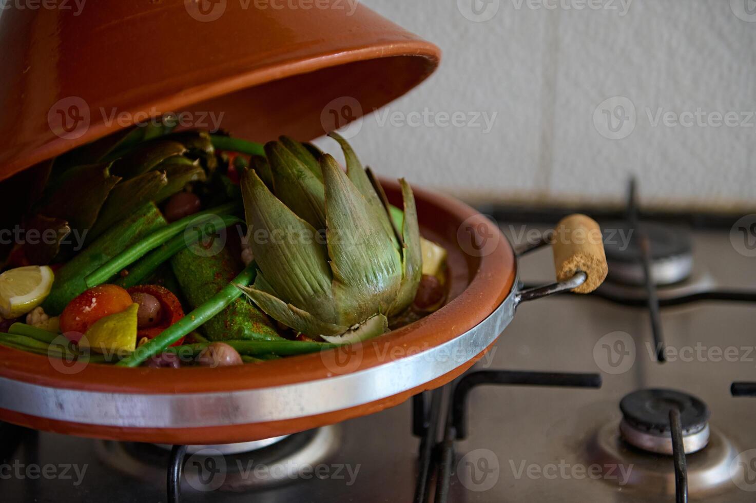 Cropped view of clay pot tagine with open loid while vegetables are steaming on the kitchen stove. Moroccan food concept photo