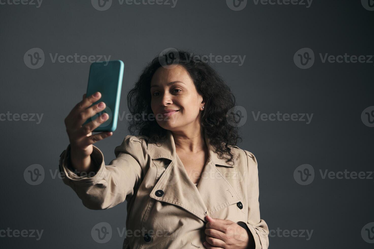 Delightful young pregnant woman in stylish beige trench, making self portrait, smiling, isolated over gray background photo