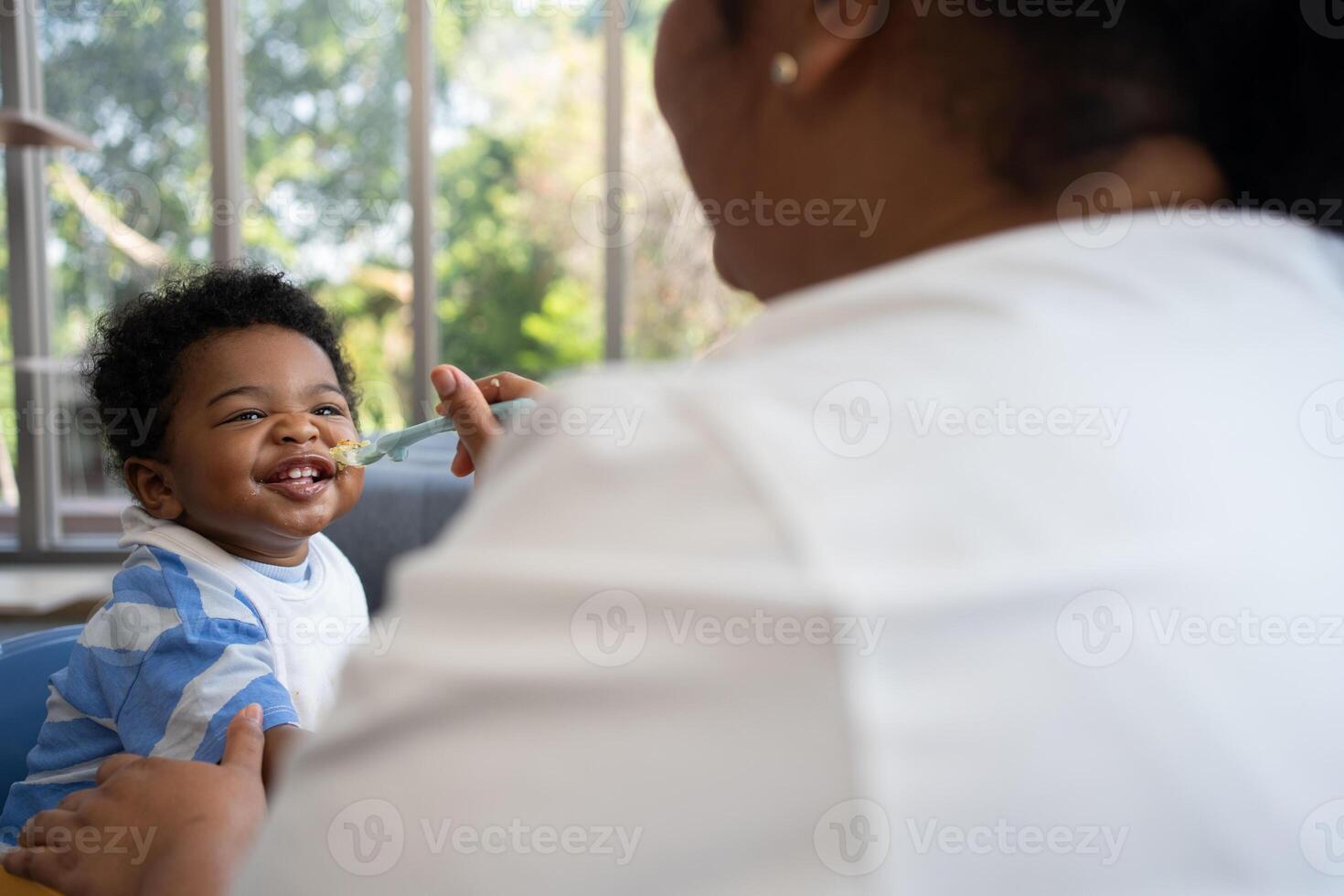 Asian mother feeding her 9 months old her cute little baby and African American helping for holding food plate At Home. Photo series of family, kids and happy people concept. Parents feed kids.