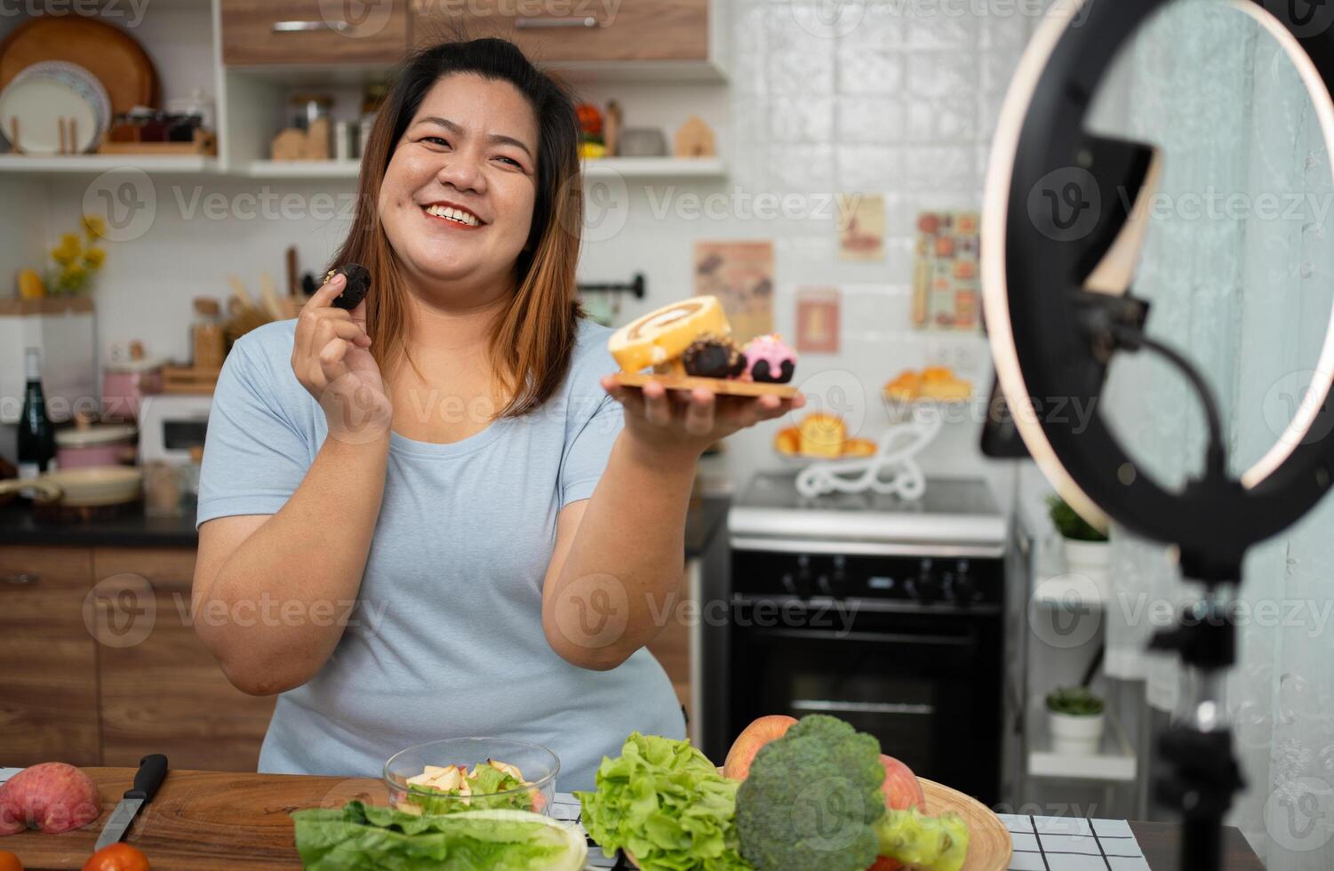 Asian Pregnant blogger looking at smartphone camera, she is live streaming cooking class for pregnant. Asian woman standing happily smiling at the kitchen counter preparing fresh organic salad. photo