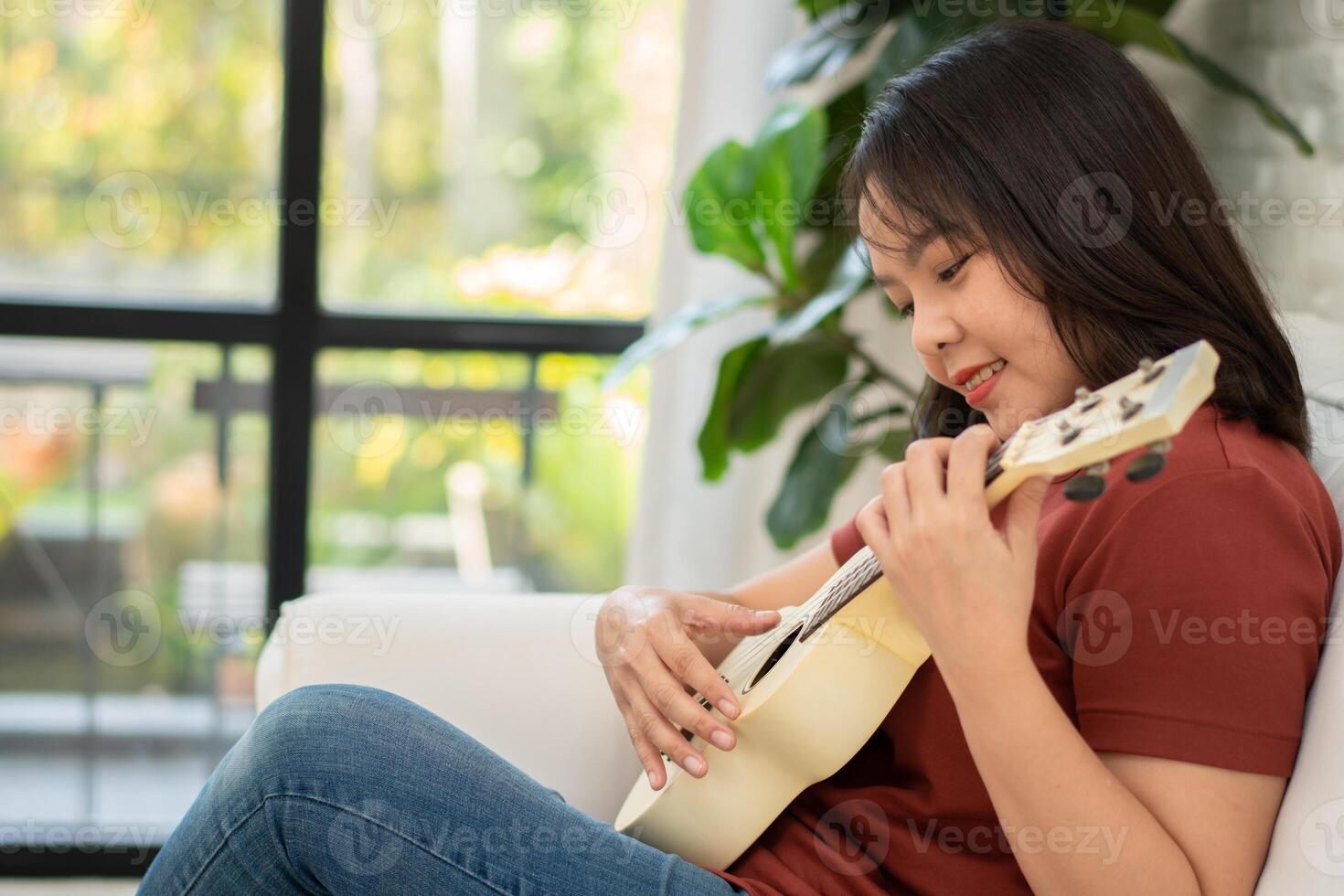 contento asiático mujer sentado en sofá en vivo habitación y practicando ukelele en ocio. relajación y inspiración desde artista. concepto de música, relajarse y pasatiempo actividad disfrutar en ocio foto