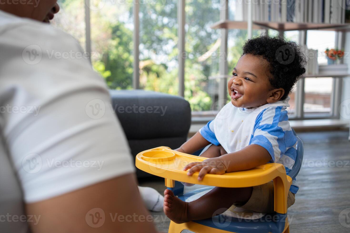 Asian mother feeding her 9 months old her cute little baby and African American helping for holding food plate At Home. Photo series of family, kids and happy people concept. Parents feed kids.