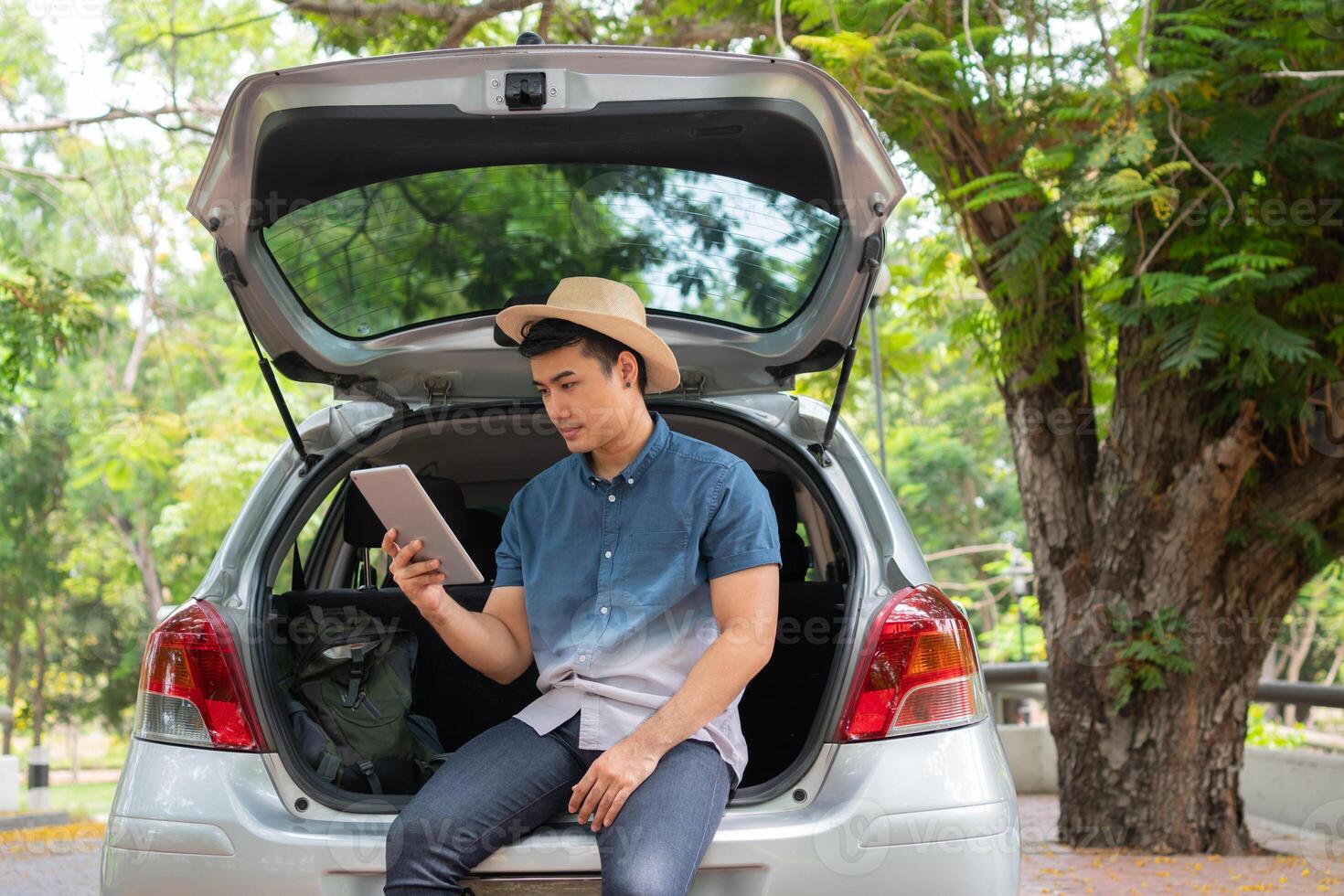 Asian man sitting back of car and holding tables check location and find place to travel. Happy road trip and vacation in summer in the countryside. Concept of journey on leisure, road trip vacation photo