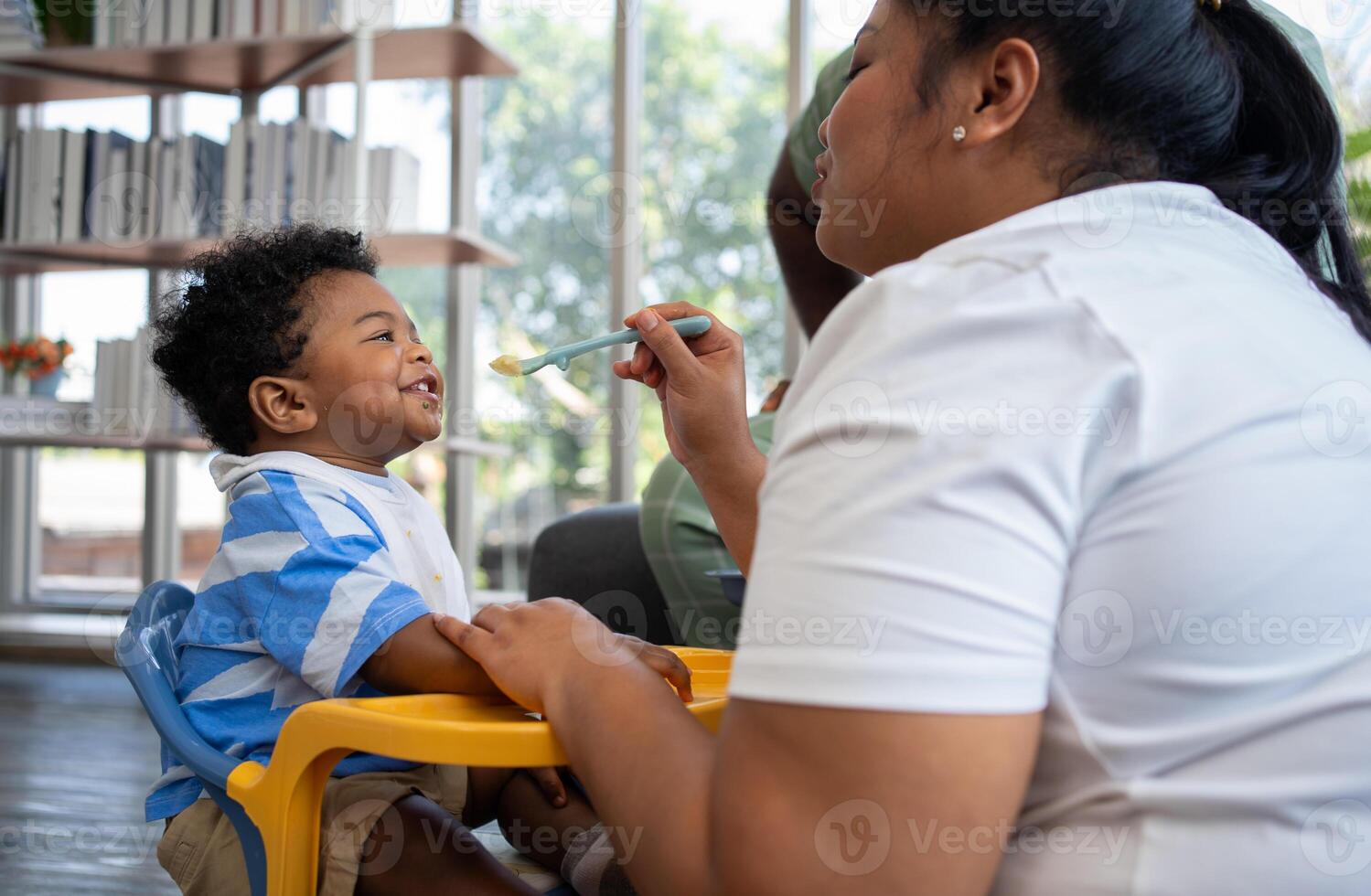 Asian mother feeding her 9 months old her cute little baby and African American helping for holding food plate At Home. Photo series of family, kids and happy people concept. Parents feed kids.